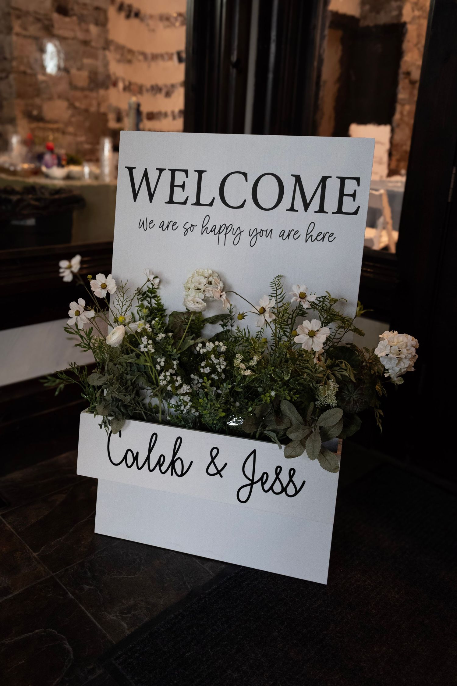 Three elegant white wedding welcome signs decorated with fresh greenery and florals against a rustic brick backdrop.