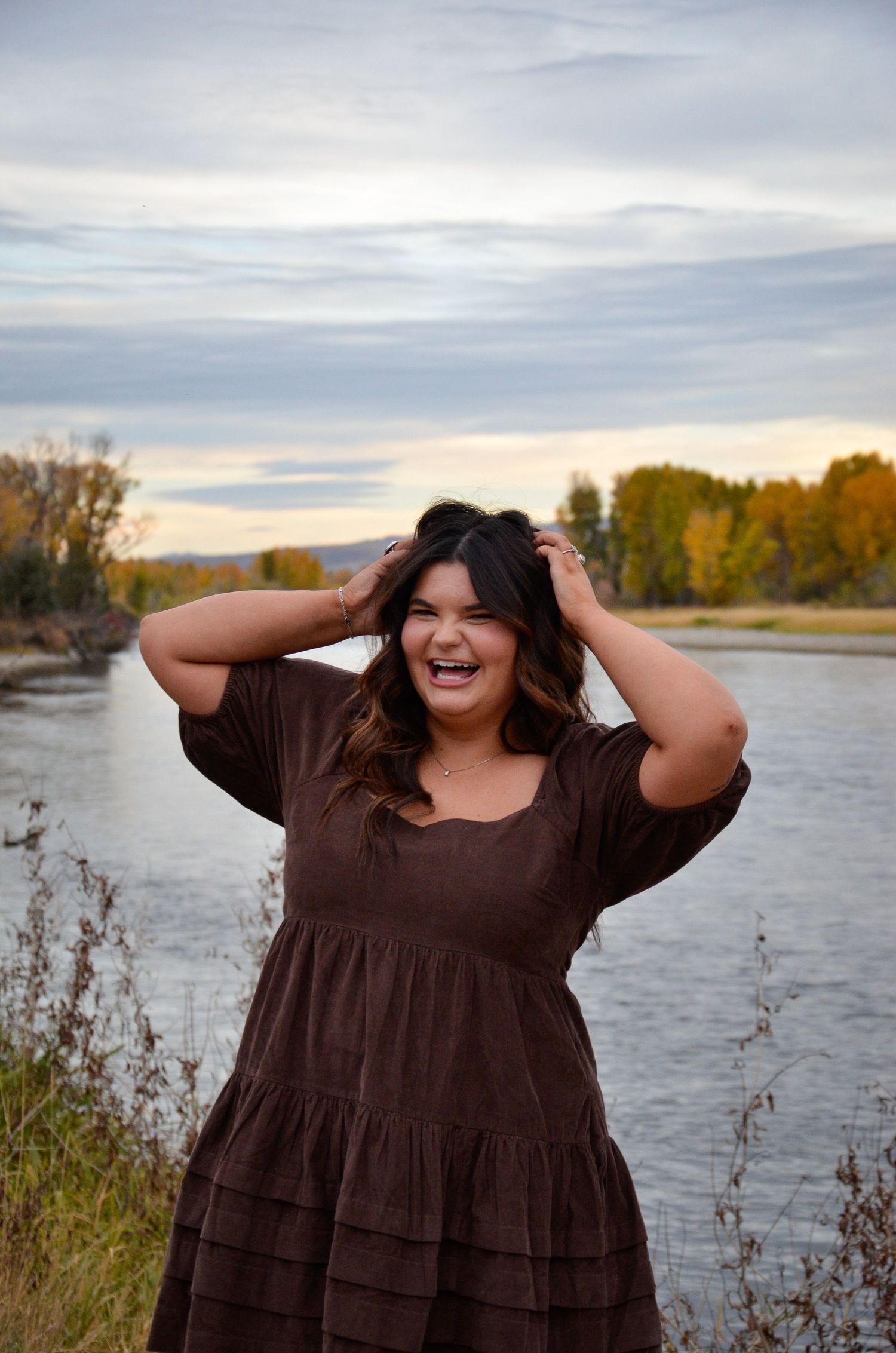 Person in brown dress laughs joyfully by riverside during golden hour.