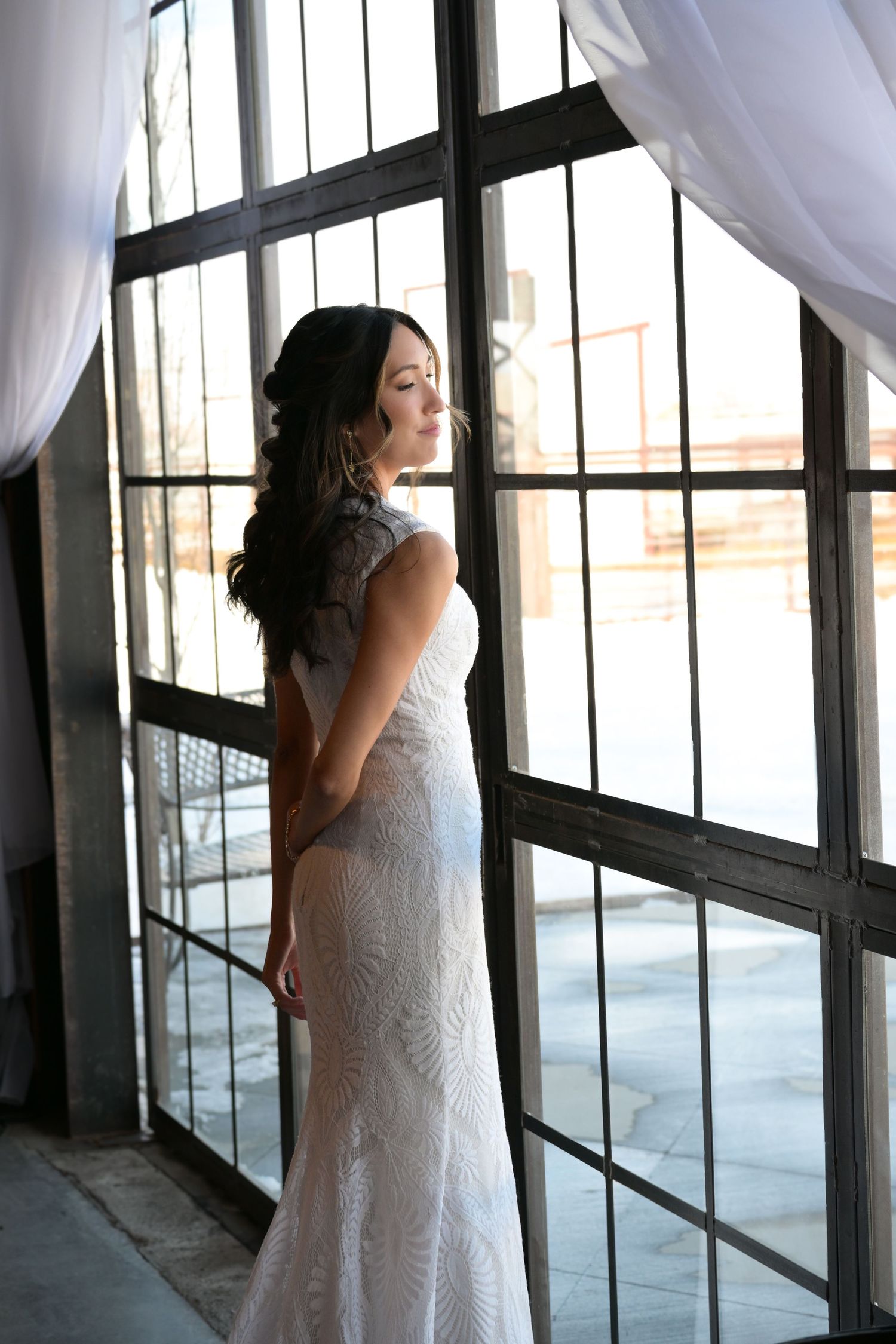 Bride in lace wedding dress poses elegantly by industrial window with natural backlighting.
