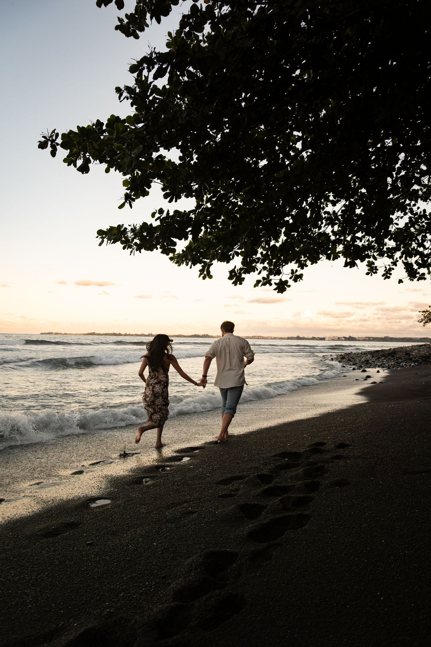 Two silhouettes walk together along a dark sandy beach at sunset with ocean waves and tree branches framing the scene.