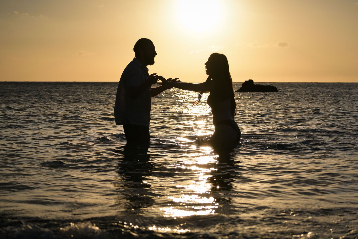 Silhouetted couple stands in ocean water at sunset creating romantic moment against golden horizon.