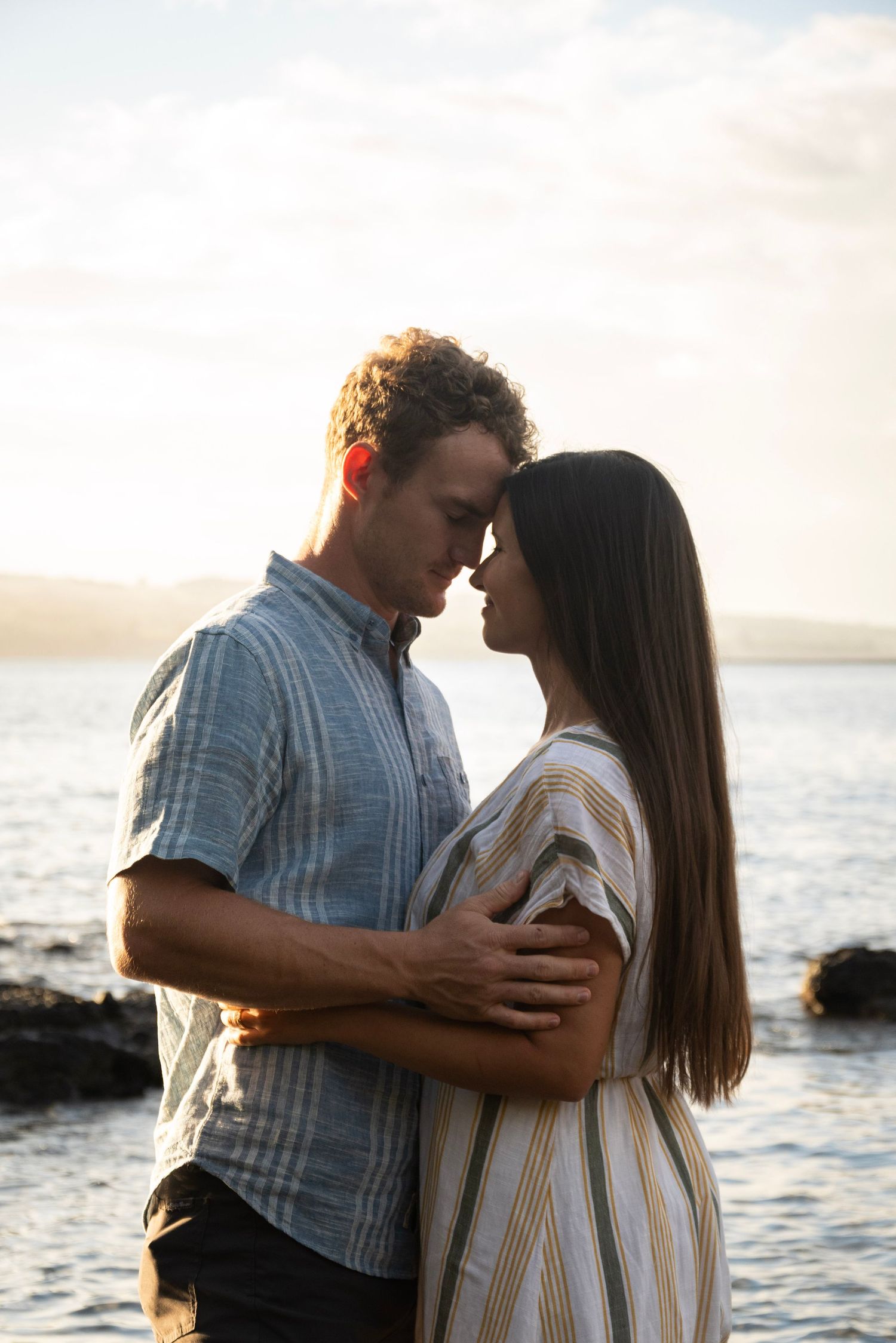 Romantic couple embrace at sunset by the ocean during golden hour as waves crash against rocks in the background.
