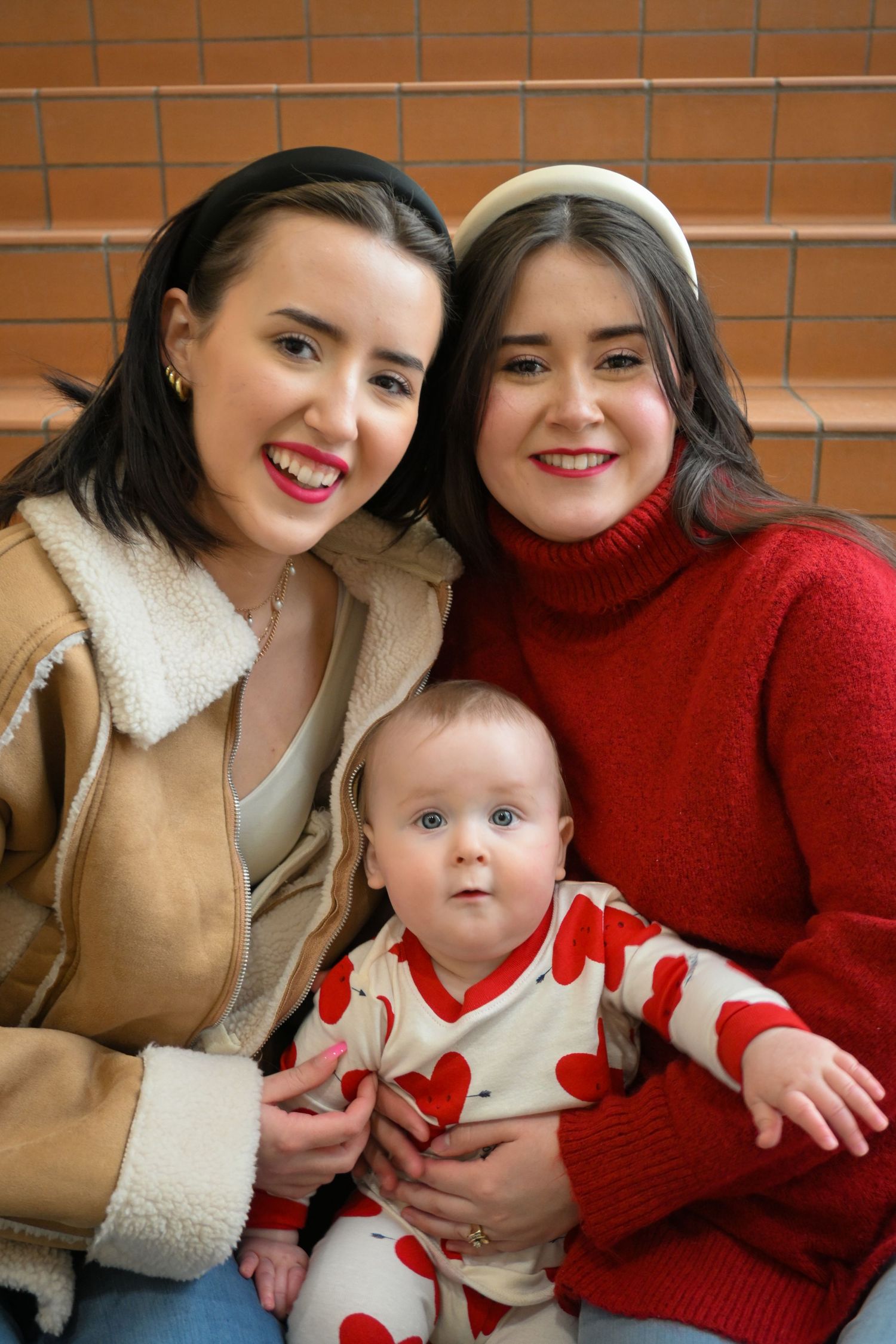 Two friends in cozy winter sweaters sitting together with an infant wearing a heart-print outfit.