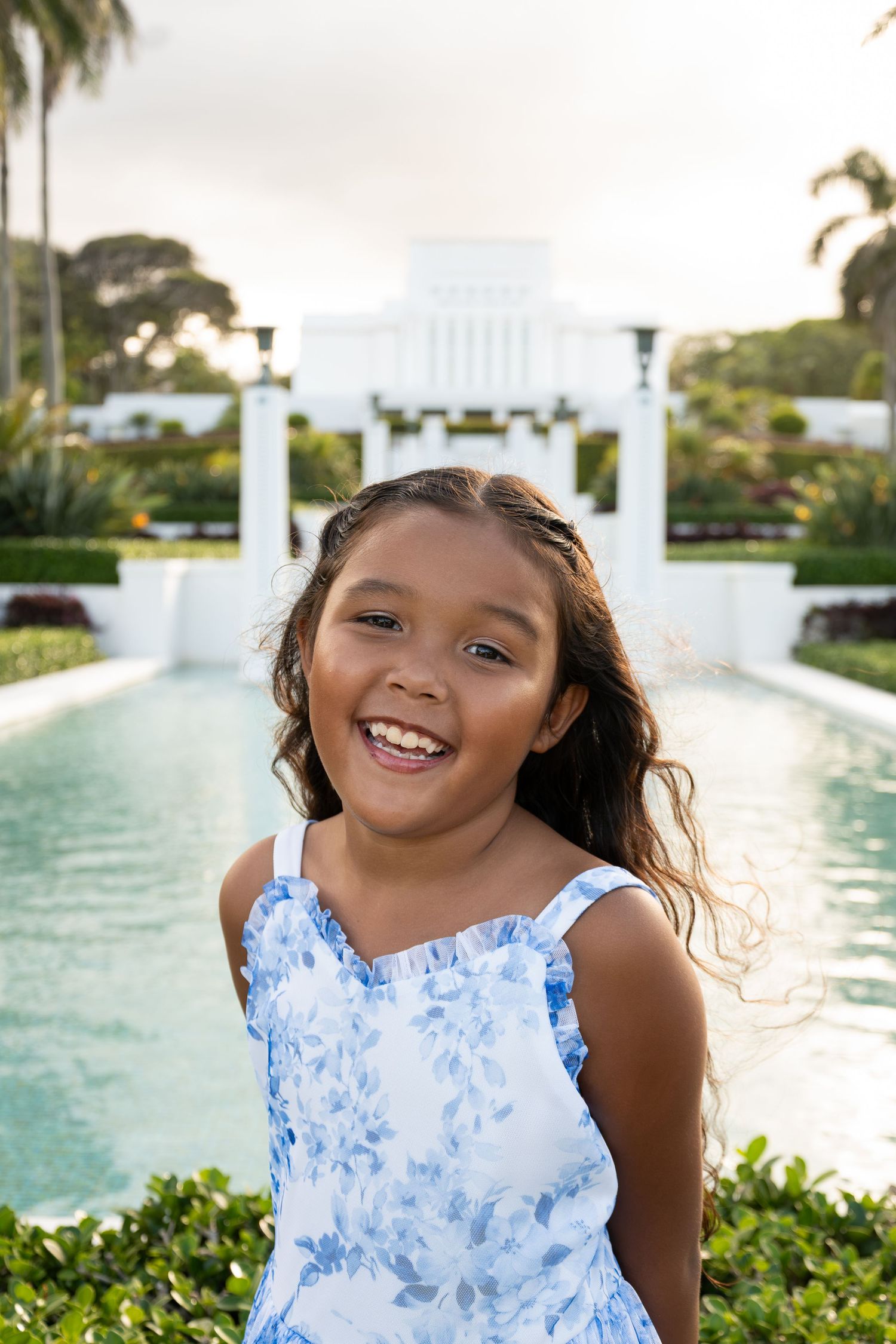 Smiling young girl in blue floral dress poses by a reflective pool at a white Hawaiian temple.