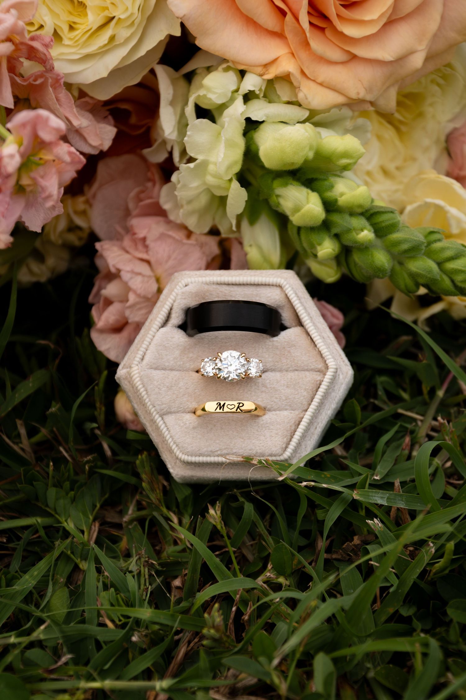 Wedding rings displayed in hexagonal velvet box beside bridal bouquet of pink and cream flowers.