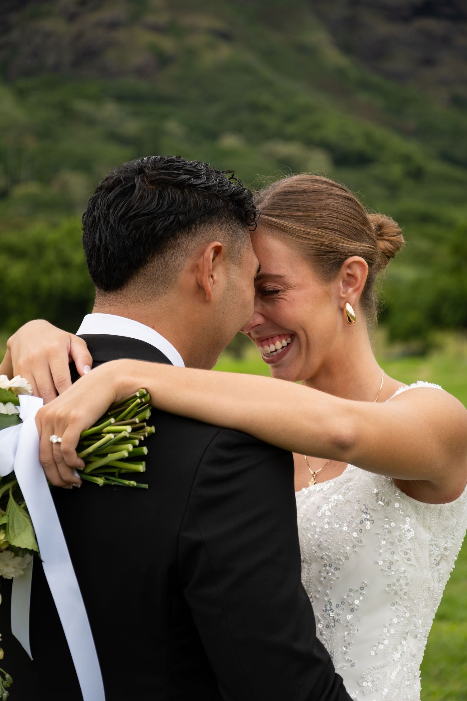 Joyful bride in white sequin gown embracing groom in black suit, laughing together with lush green mountains behind.