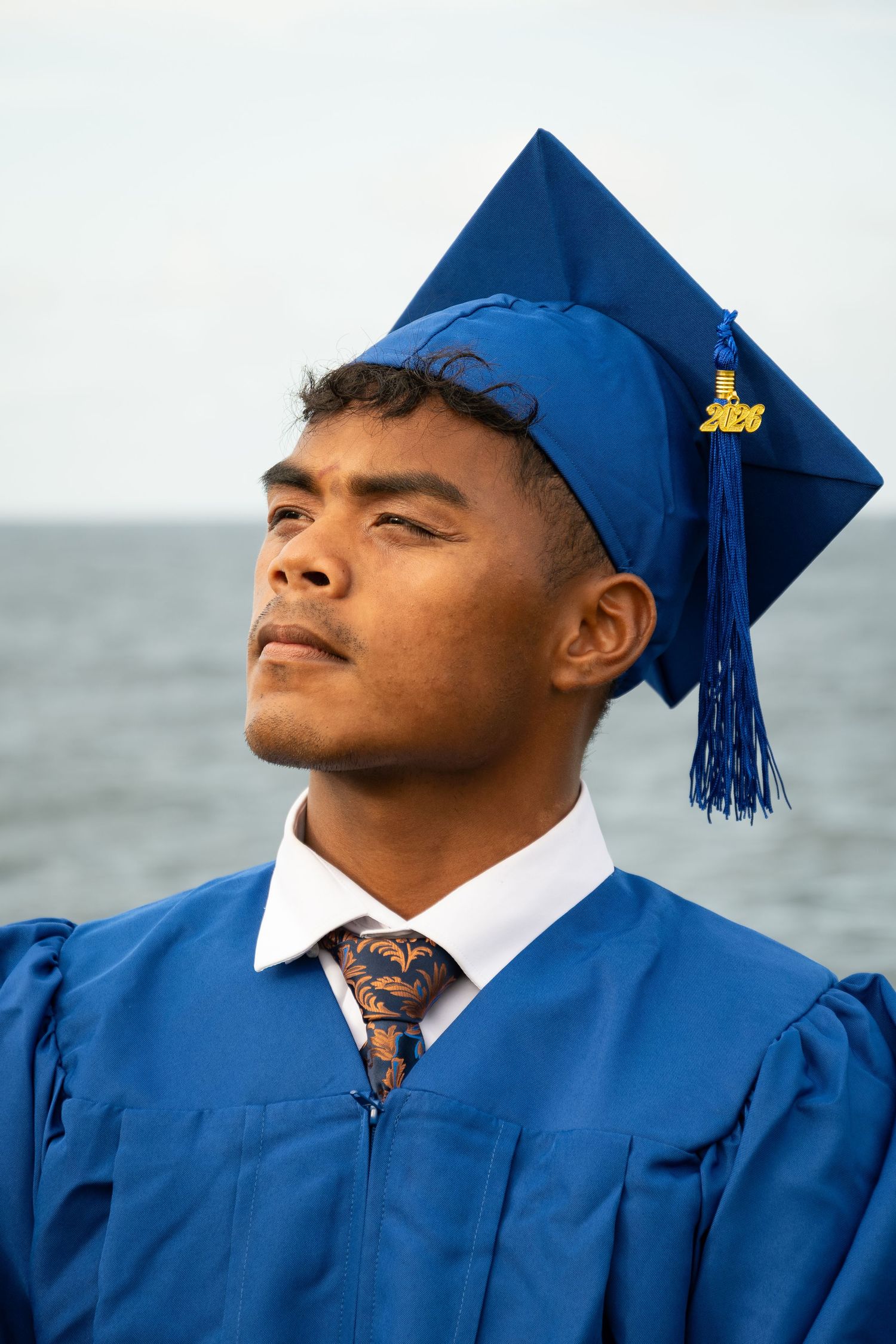 Young man in blue graduation cap and gown with tassel, posing outdoors near ocean, looking upward confidently.