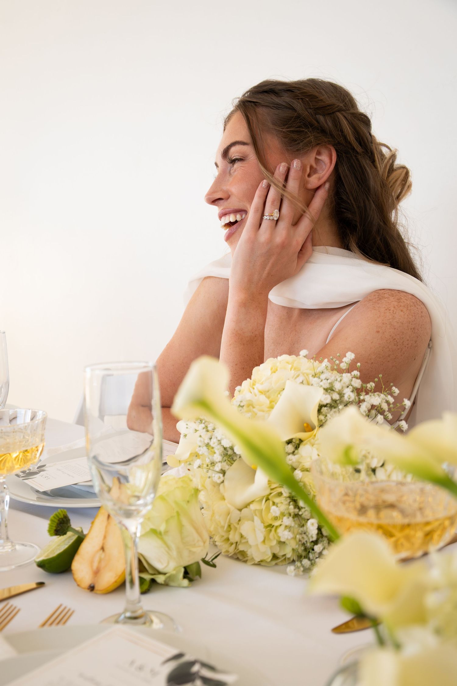 Smiling bride in white dress laughing at elegant wedding reception table with white floral centerpiece.