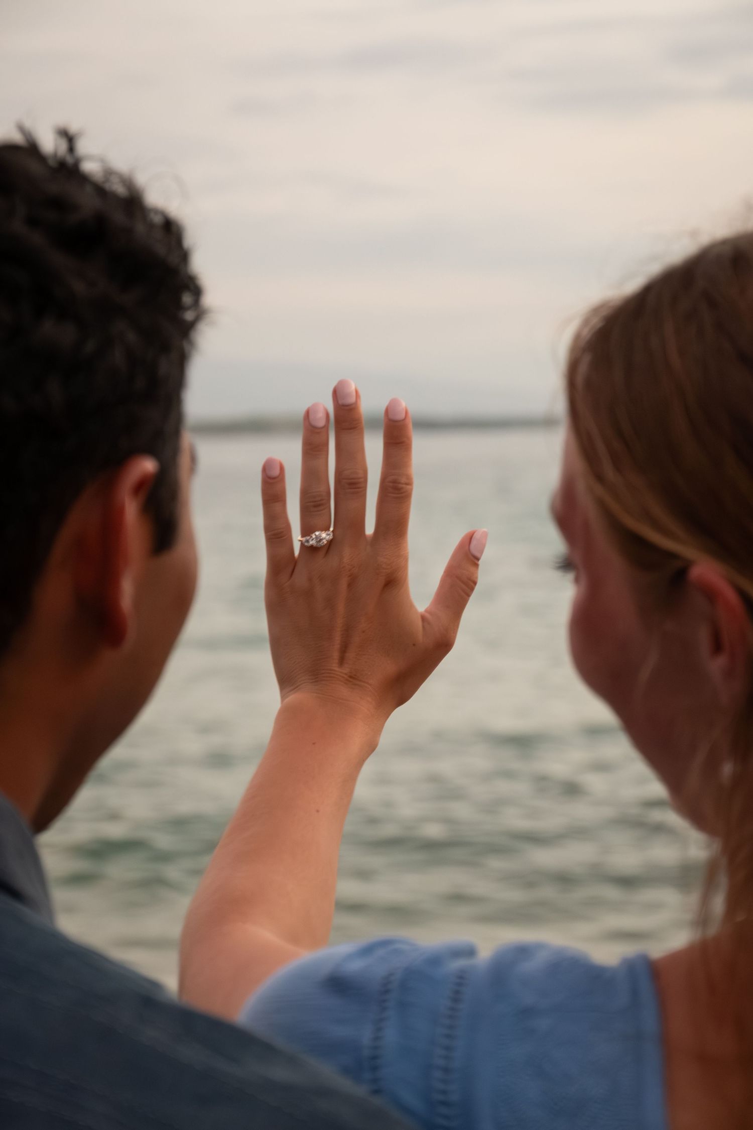 Couple showing off engagement ring with diamond solitaire by the ocean, viewed from behind.