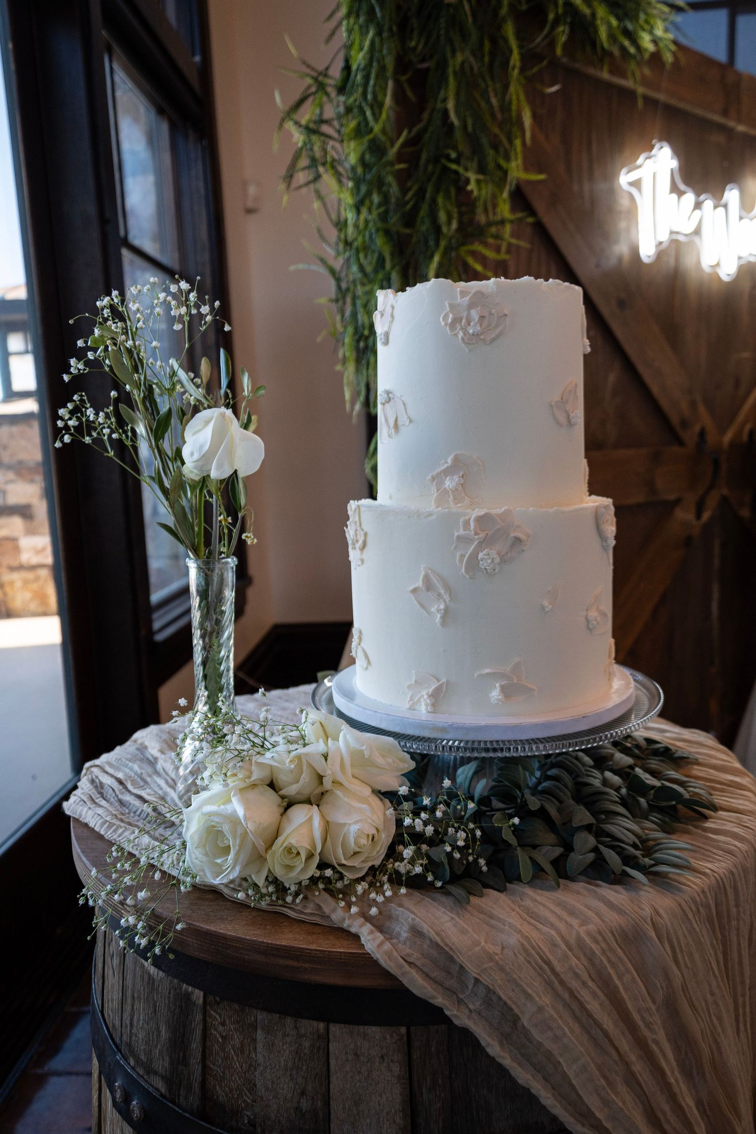 A white two-tier wedding cake decorated with delicate floral details sits on a rustic barrel with fresh roses and greenery.