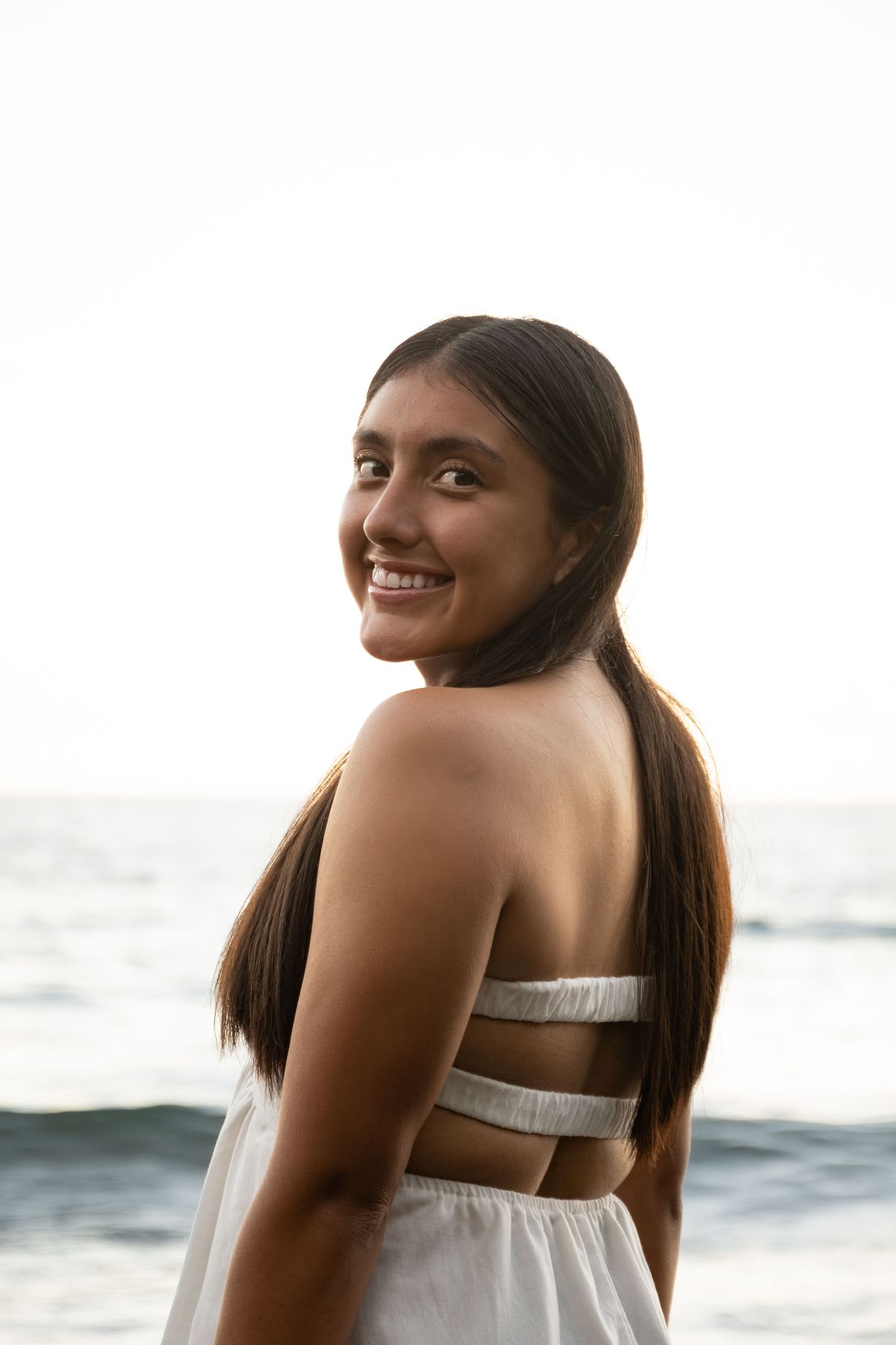 A person in a white dress smiles over their shoulder while posing at the beach during sunset.