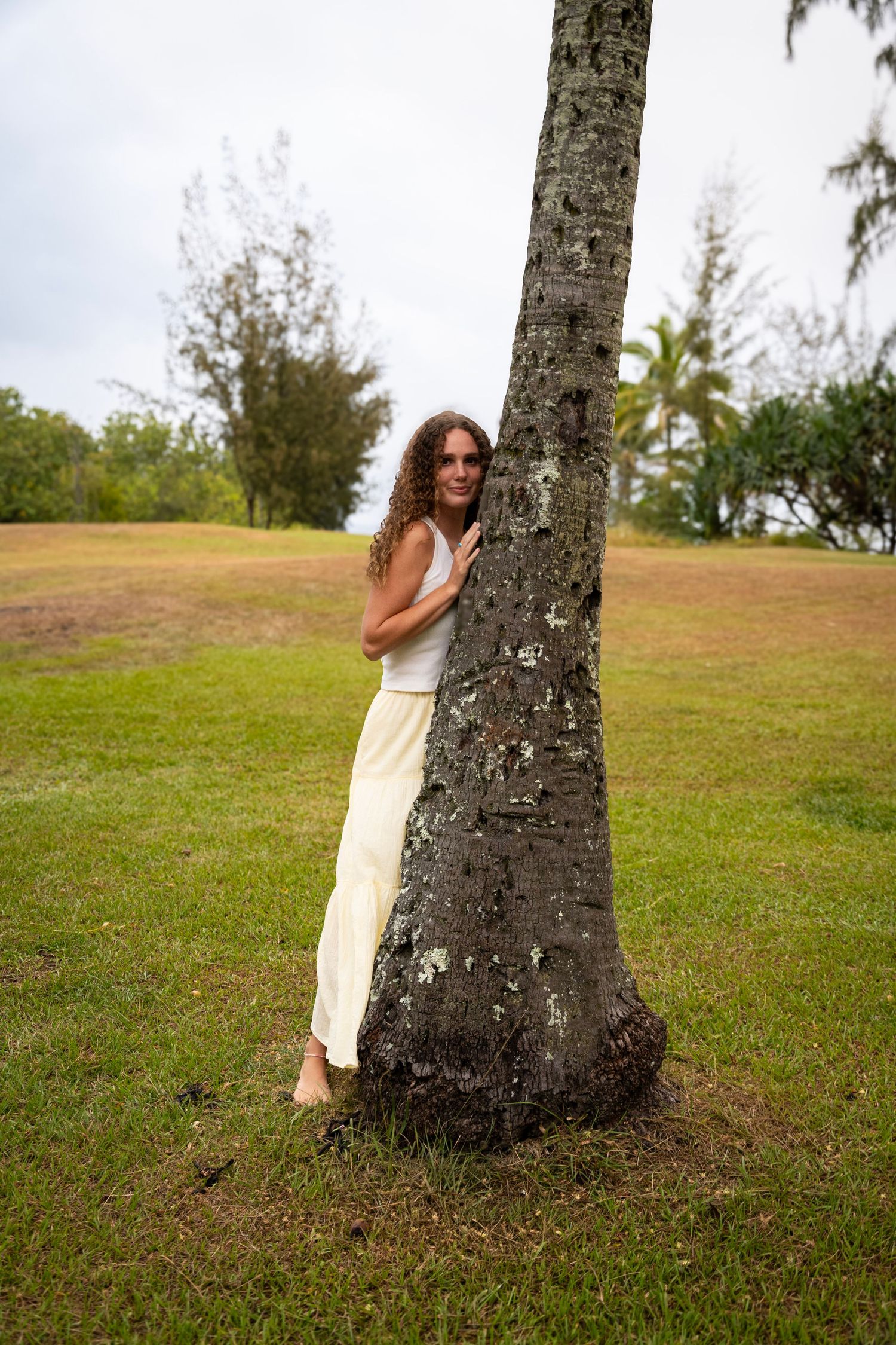Person in white outfit poses playfully behind a palm tree trunk on grassy tropical grounds.
