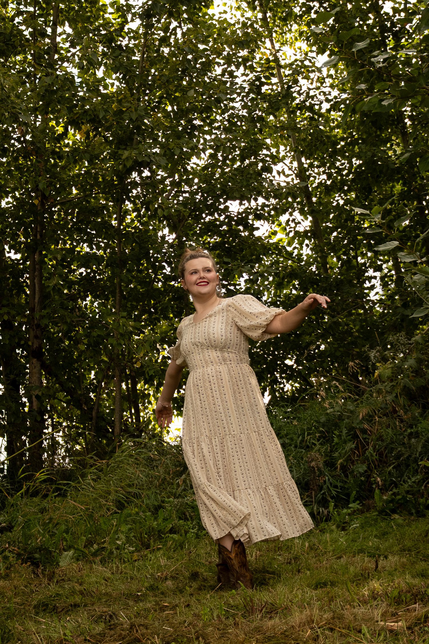 A smiling woman in a flowy cream dress poses gracefully among lush green trees outdoors.
