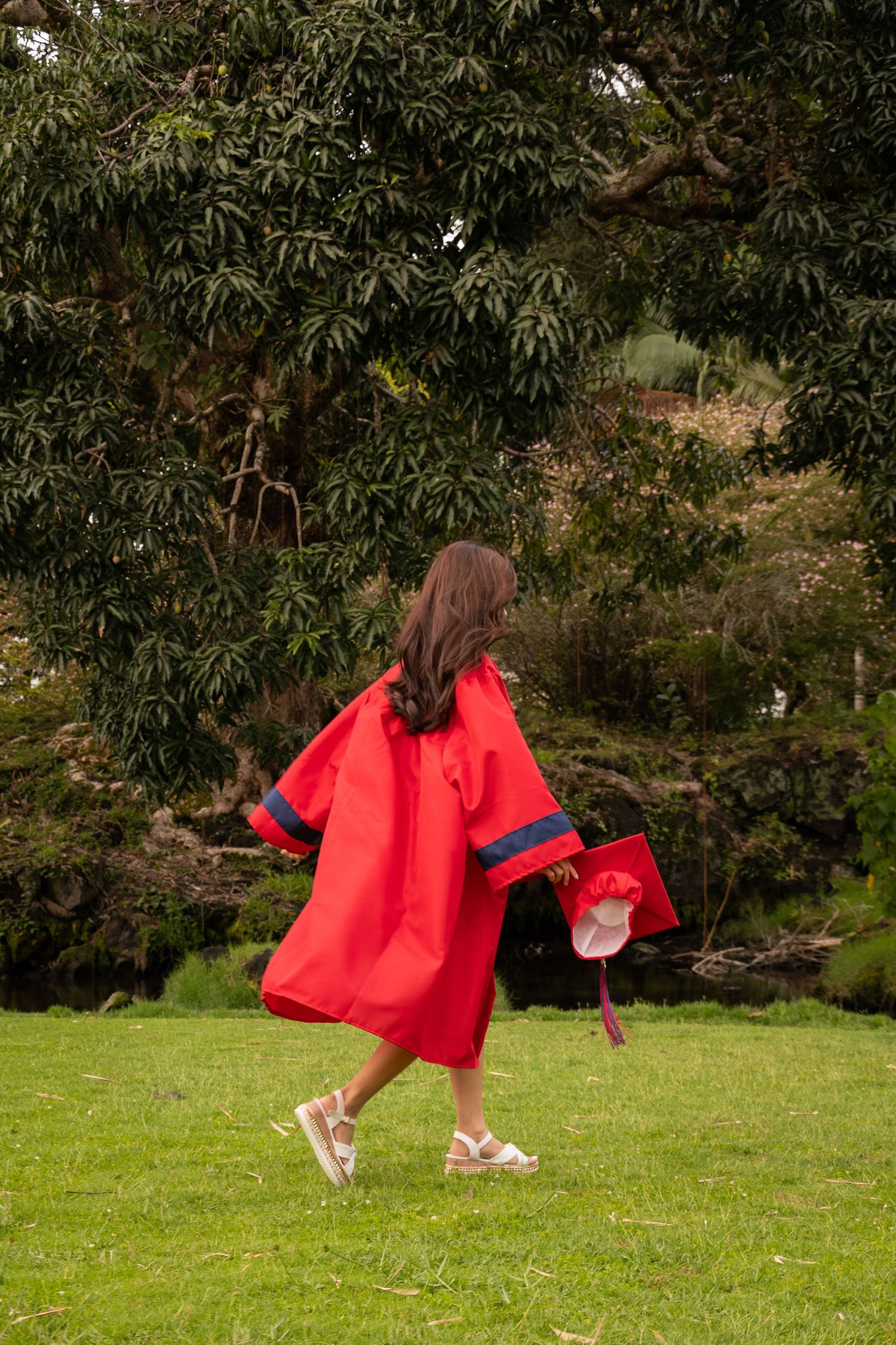 Young girl in red graduation gown and cap walking on green grass with large trees in background.