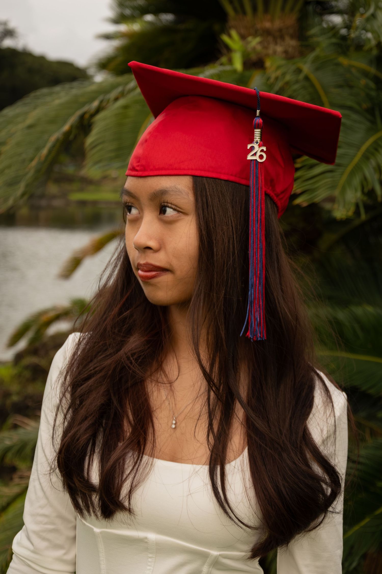 Young woman in red graduation cap with '26' charm and blue tassel, wearing white dress outdoors near tropical plants.