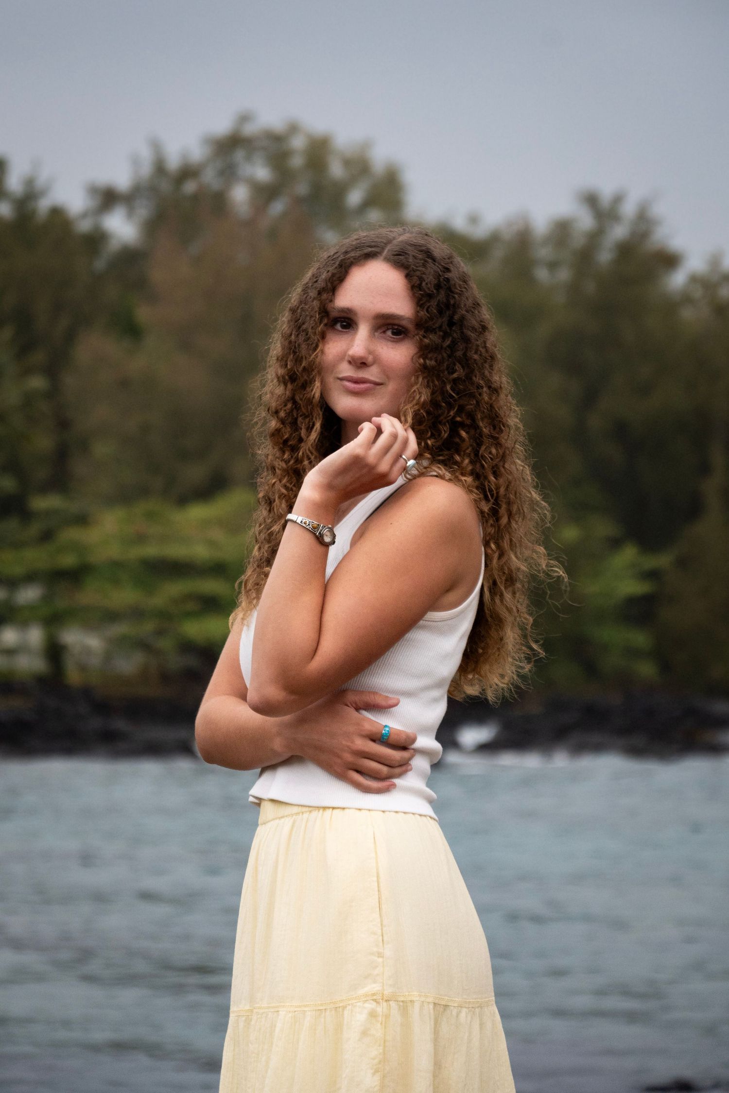 A person with long curly hair poses outdoors near a rocky coastline wearing a white top and cream-colored skirt.