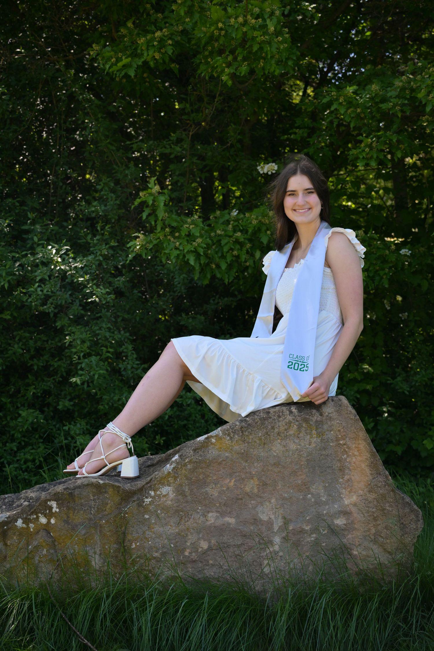 A person in a white dress shirt poses gracefully on a large rock in a natural outdoor setting with green foliage.