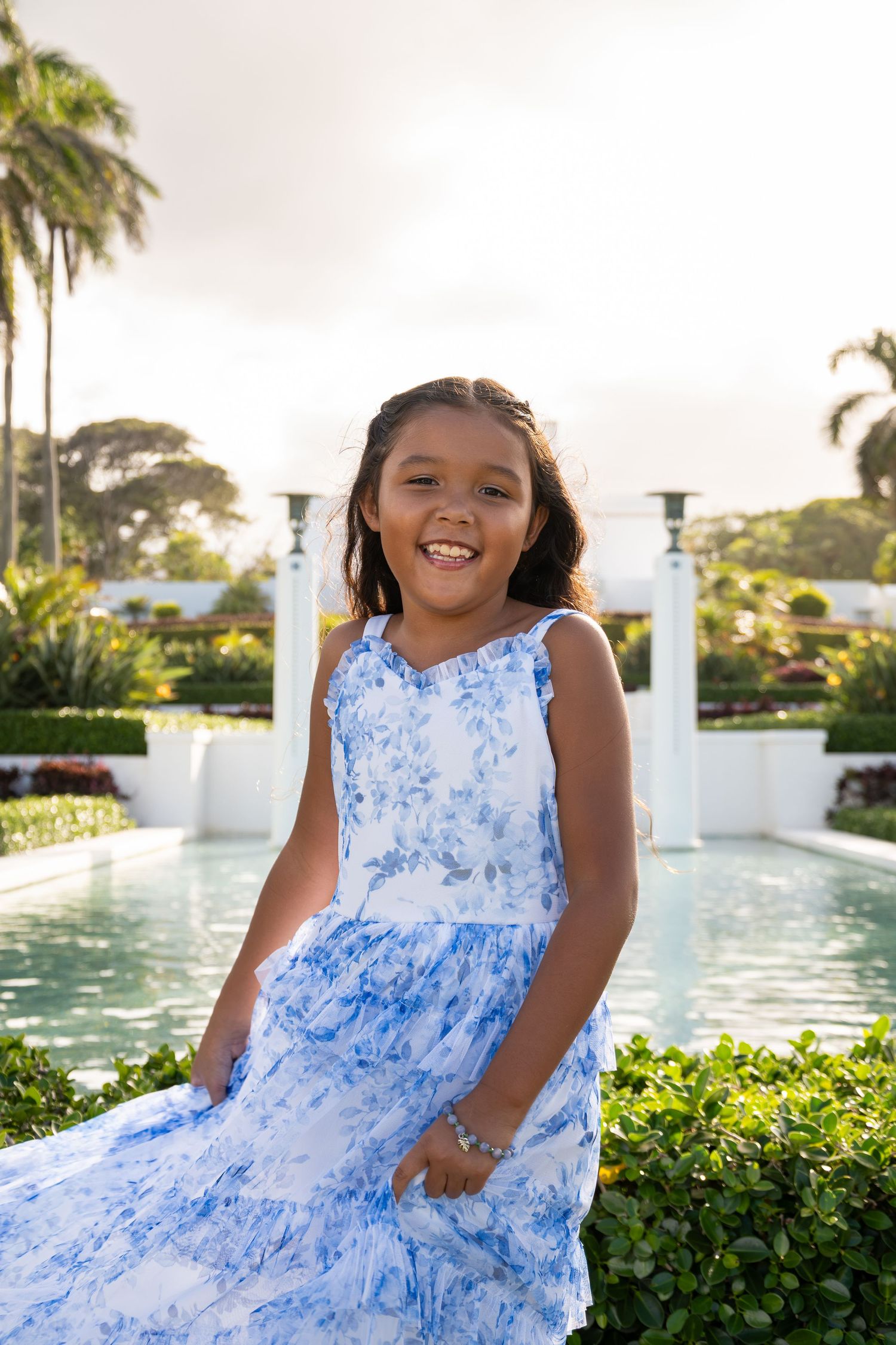 Smiling young girl in blue floral dress posing by a tropical garden pool with palm trees and white columns.