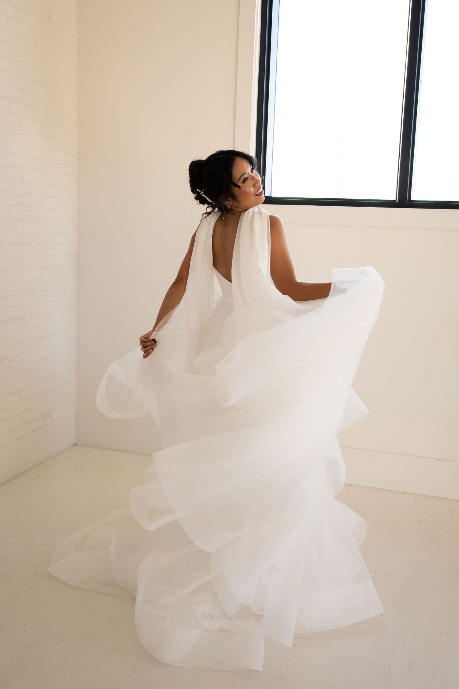 Bride in a flowing white ruffled wedding gown twirling in a bright white room near a window.