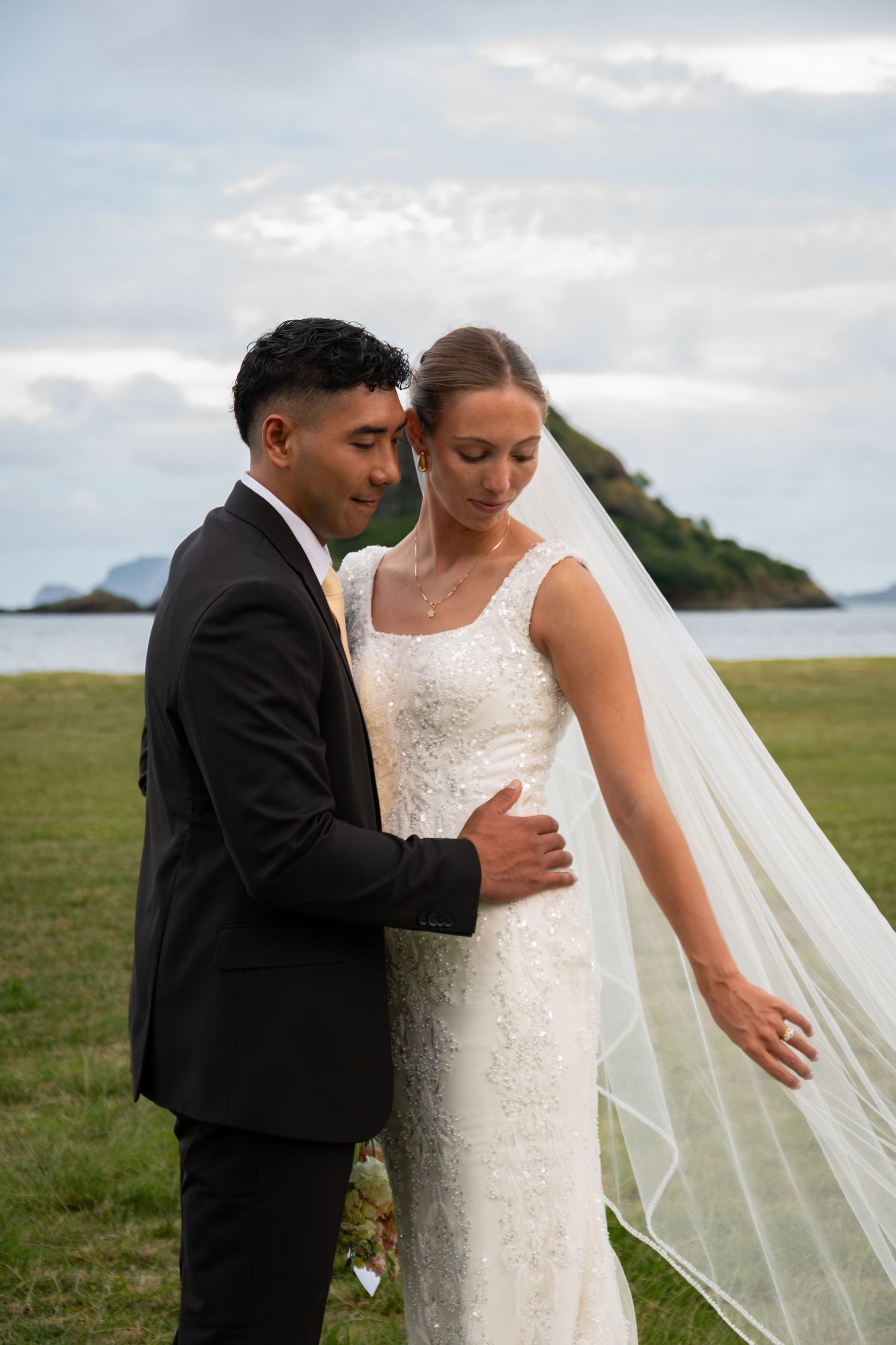 A bride and groom embrace outdoors near the ocean, she wearing a lace gown and veil, he in a black suit.