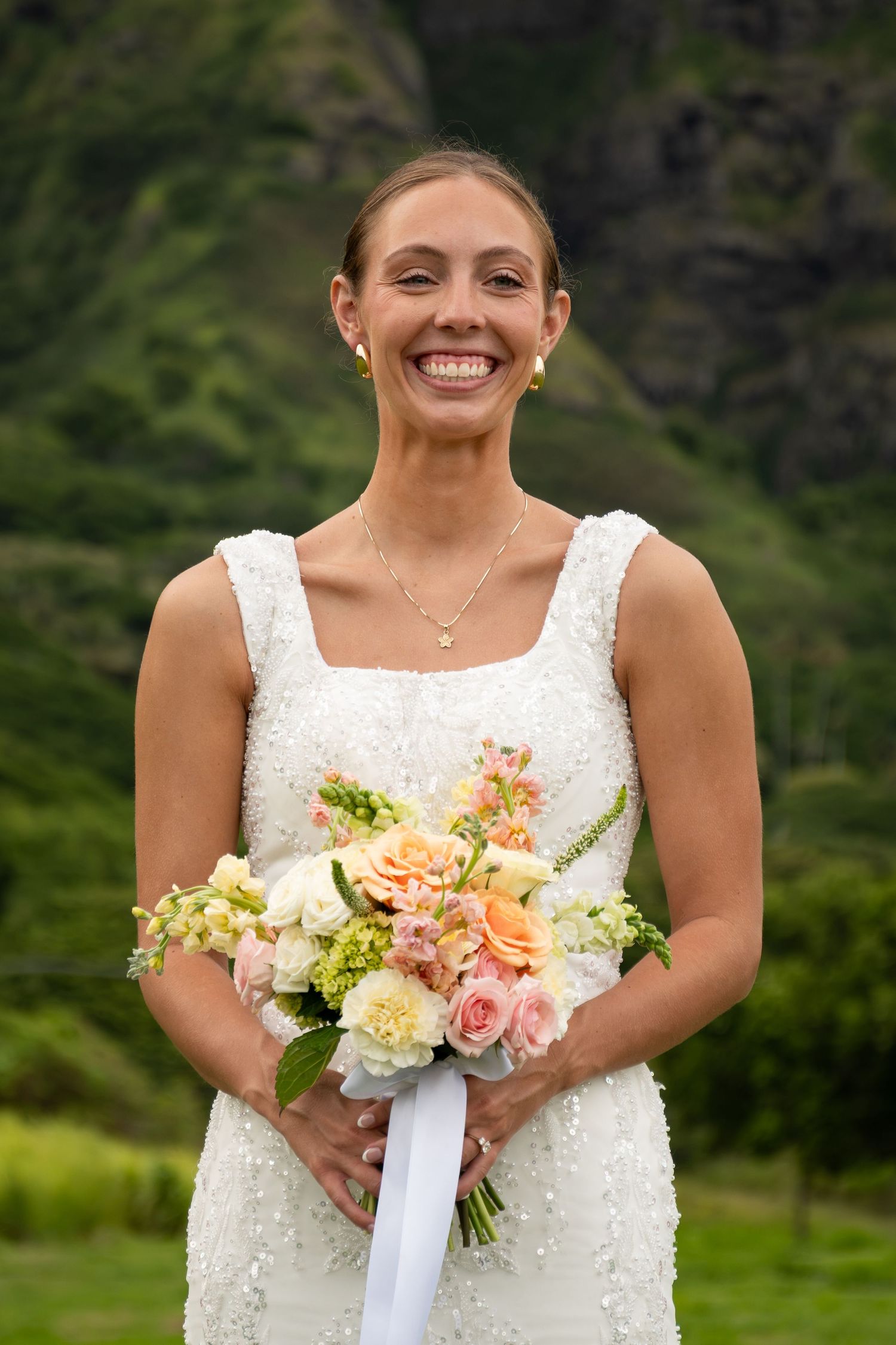 Smiling bride in white beaded gown holding peach and white floral bouquet with lush green mountains behind her.