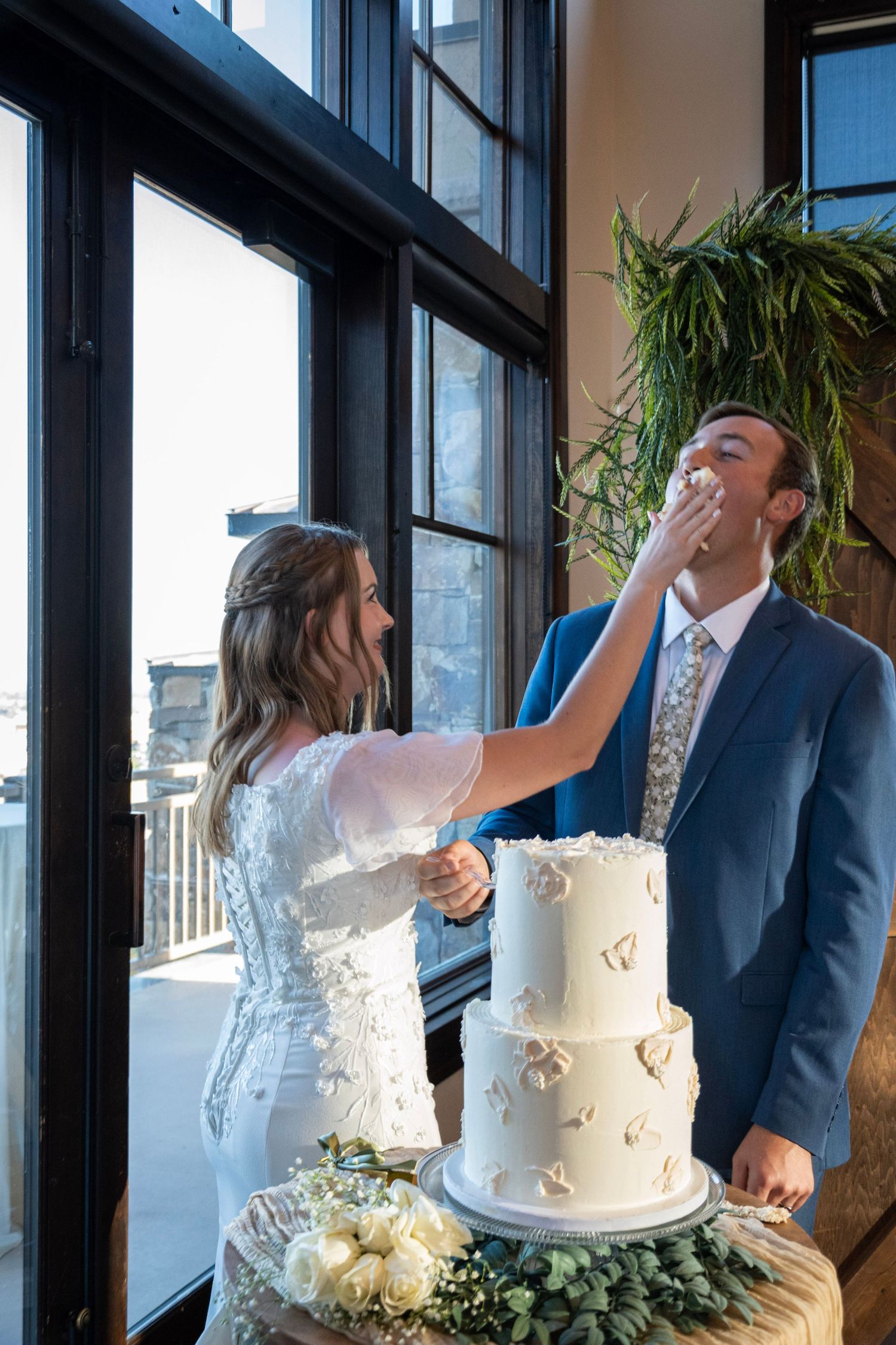 Two people share a playful moment while feeding each other wedding cake in front of large windows.