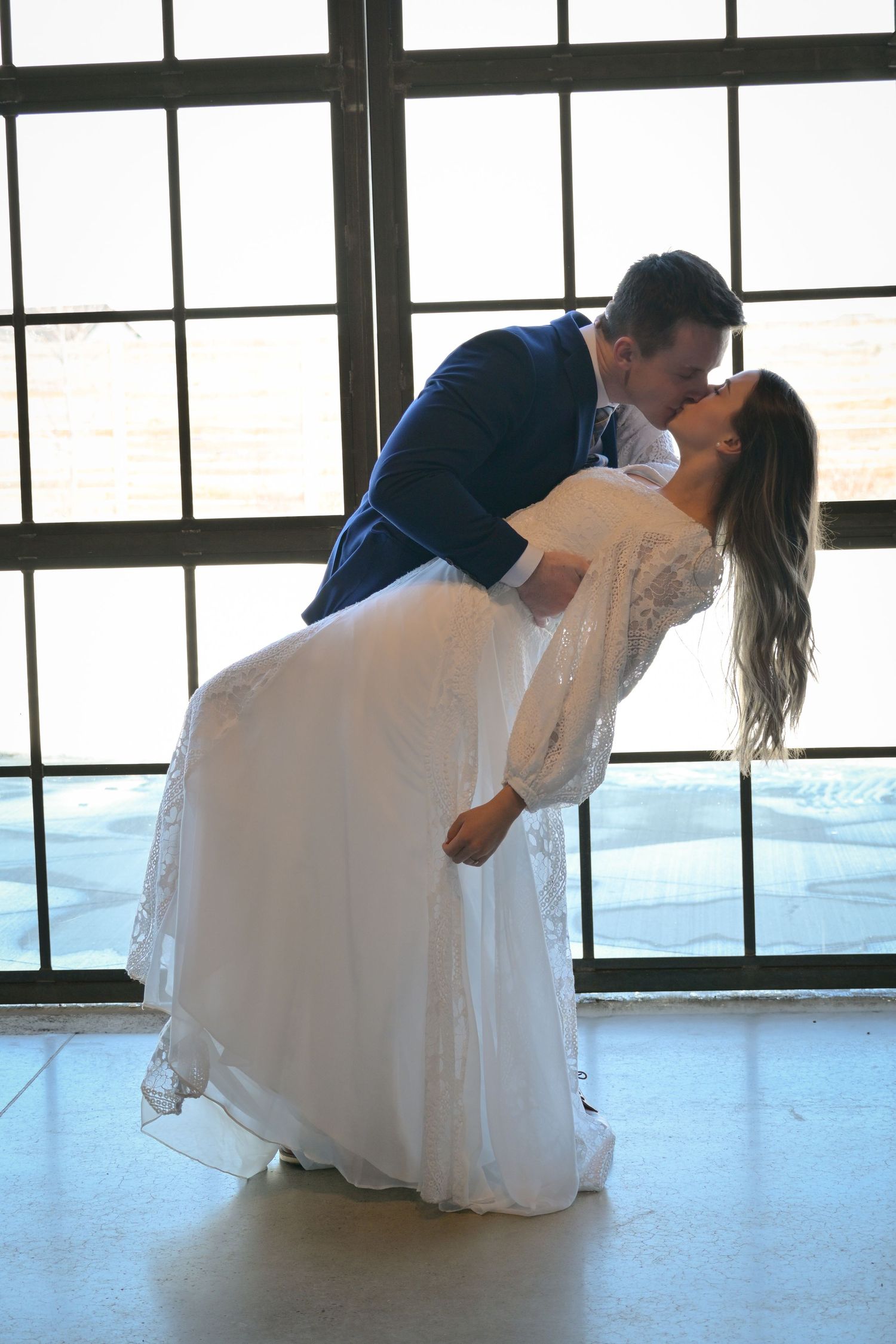 Romantic dip pose of couple by large industrial windows in wedding attire.