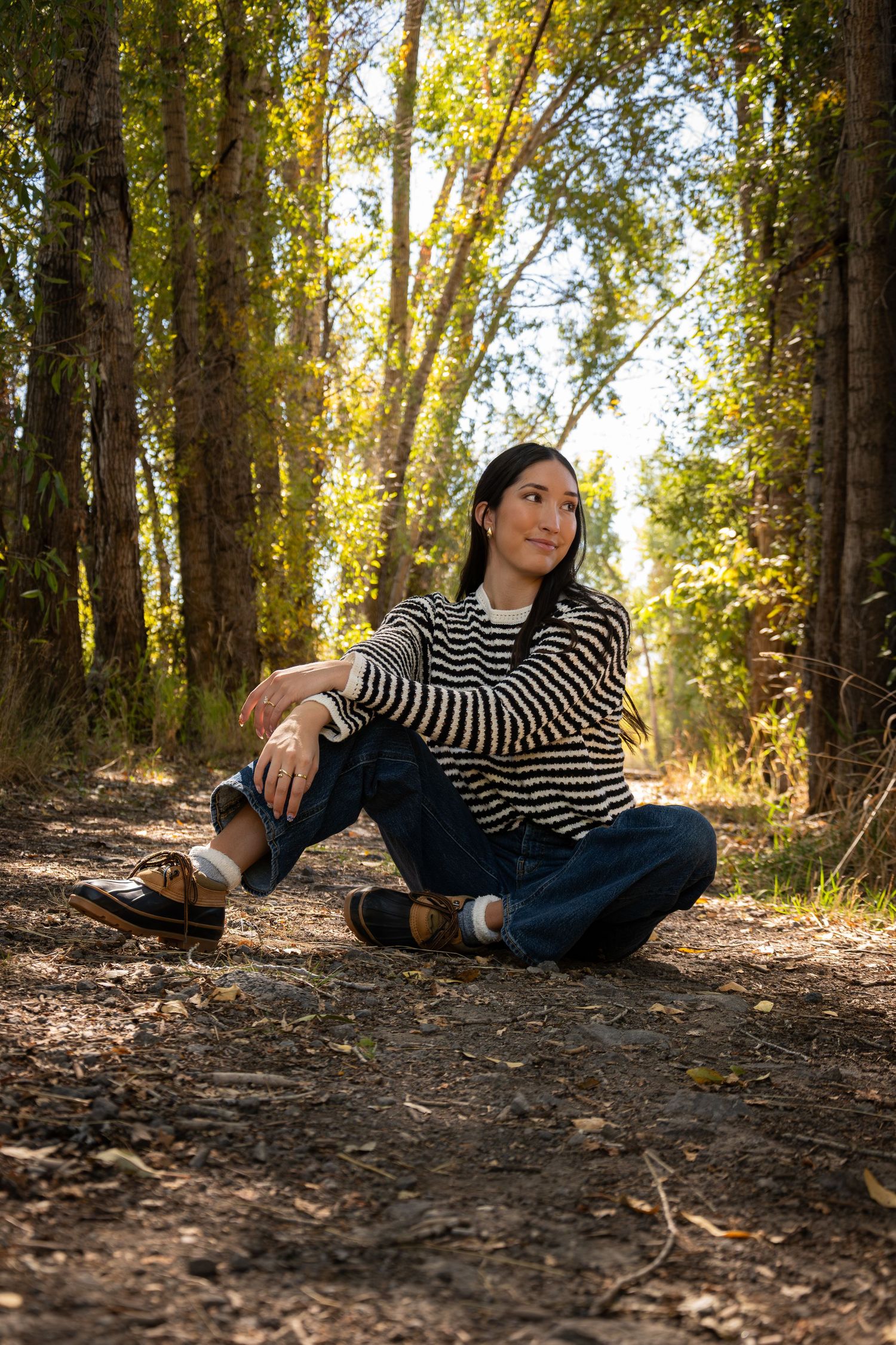 Person in striped sweater sitting on forest path with sunlight filtering through autumn trees.