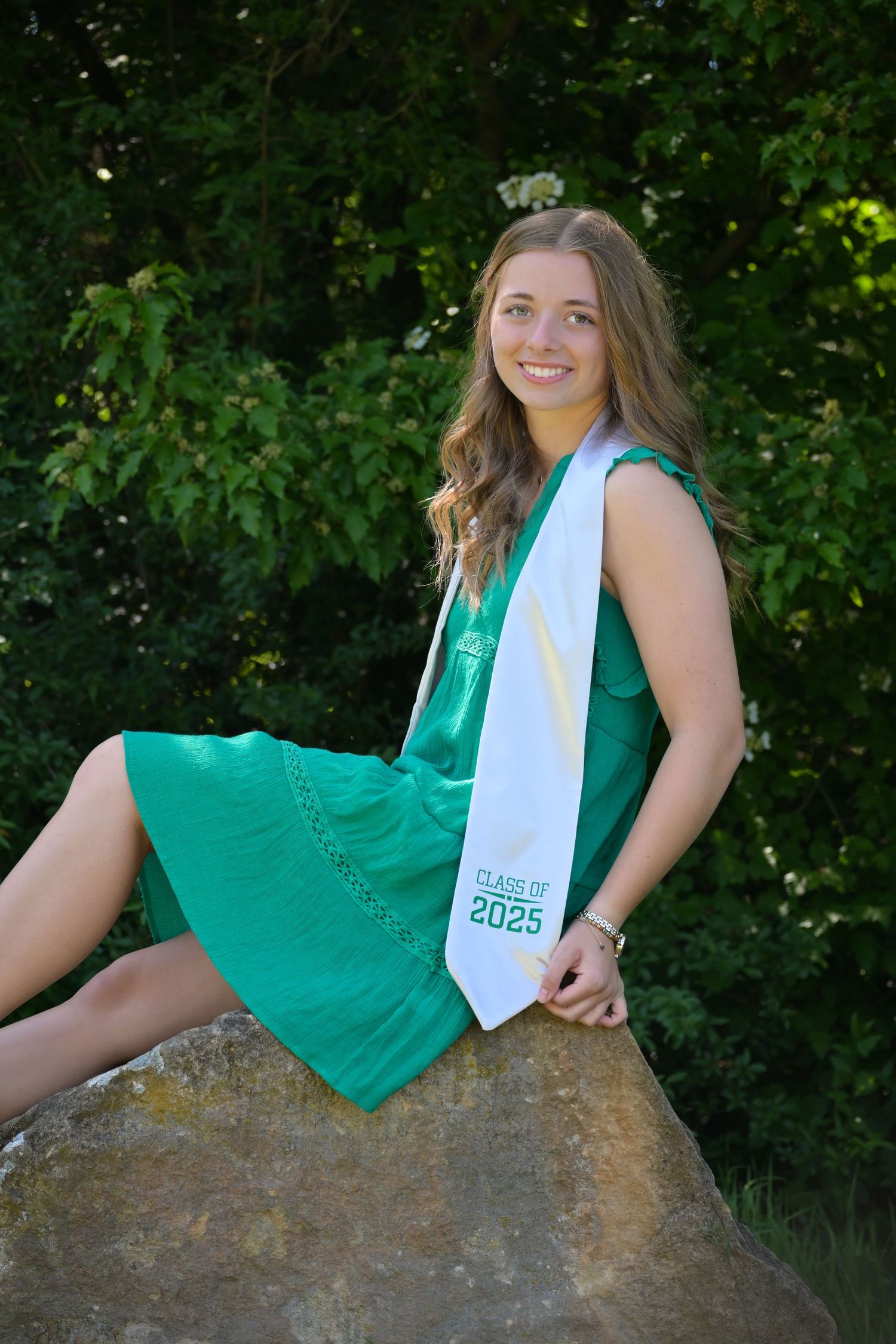 Graduate wearing emerald green dress and white sash sits on rock with natural foliage background.