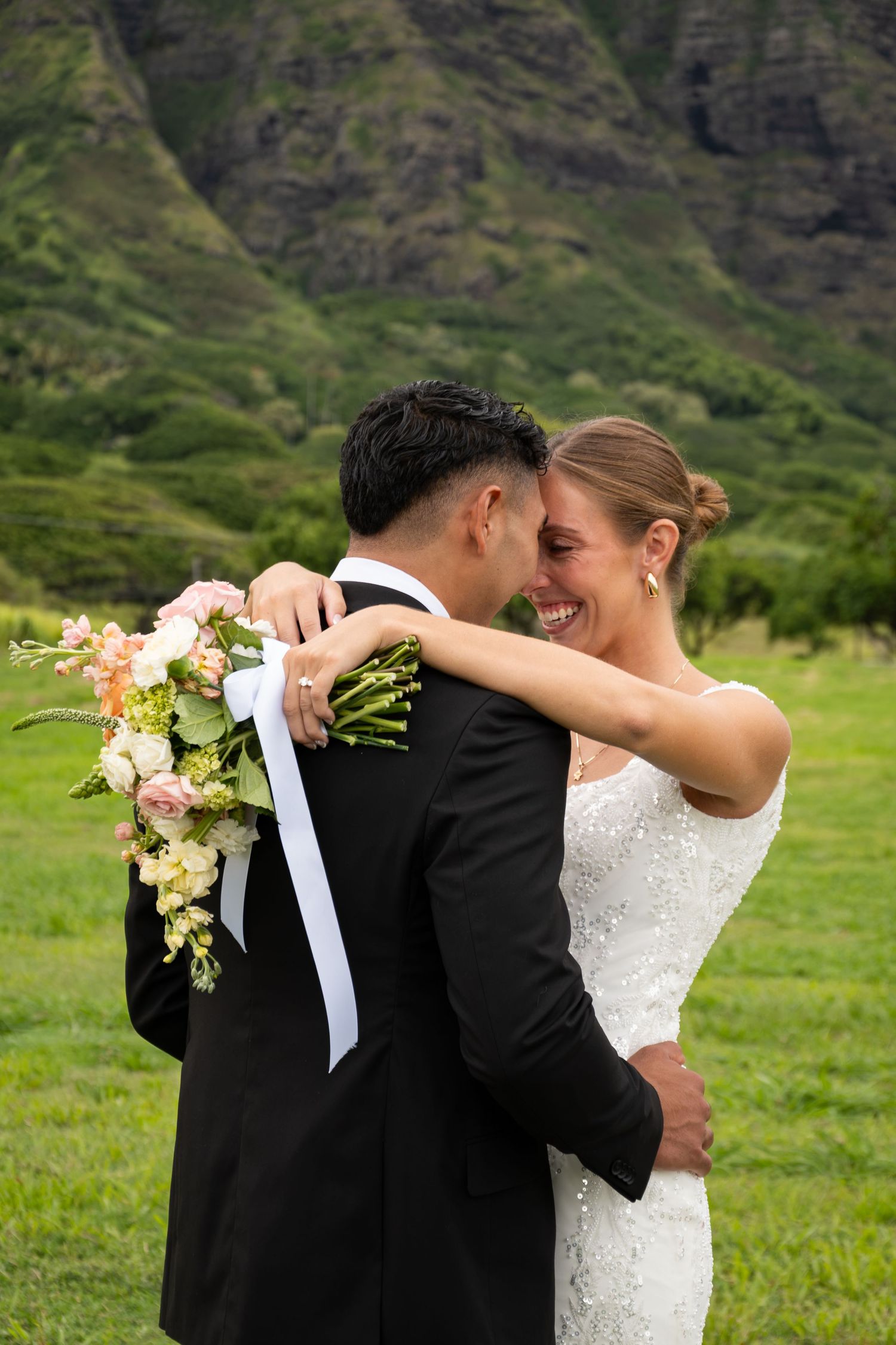A bride and groom embrace in a lush green Hawaiian valley, the bride smiling joyfully while holding a pastel floral bouquet.