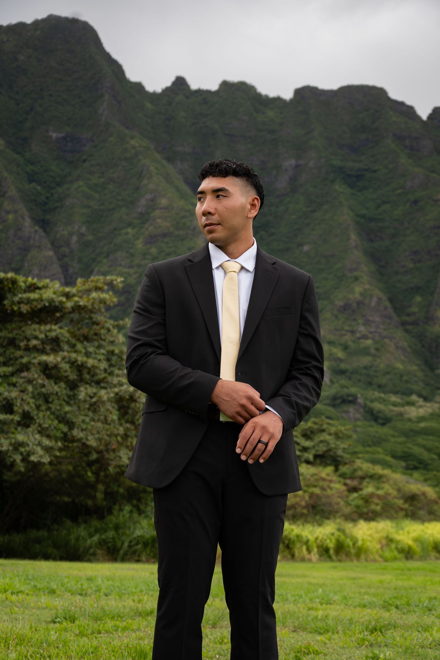 A man in a black suit and gold tie stands on green grass with lush Hawaiian mountains in the background.