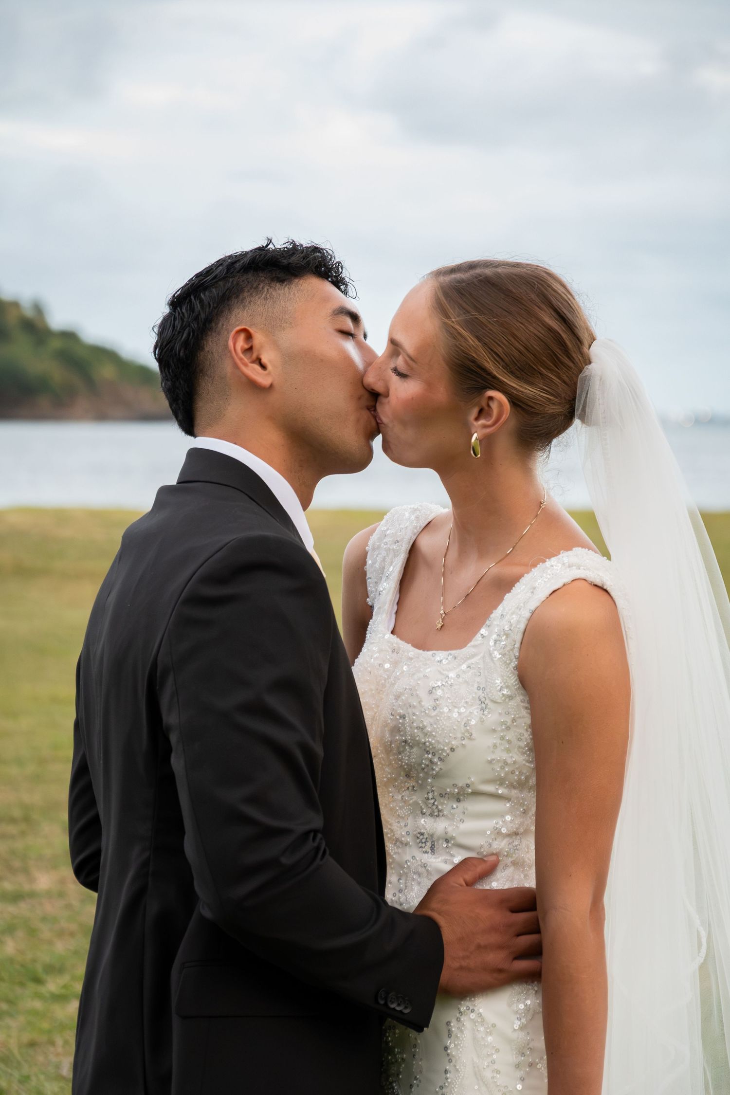 Bride and groom share first kiss outdoors in wedding attire with scenic waterfront background on their wedding day.