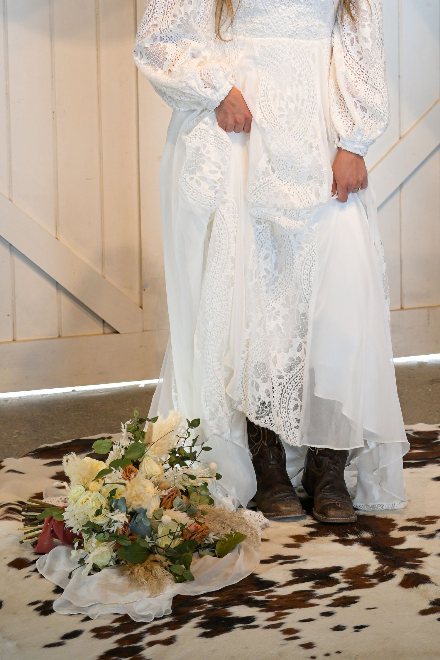 White lace wedding dress with boots and floral bouquet on cowhide rug.