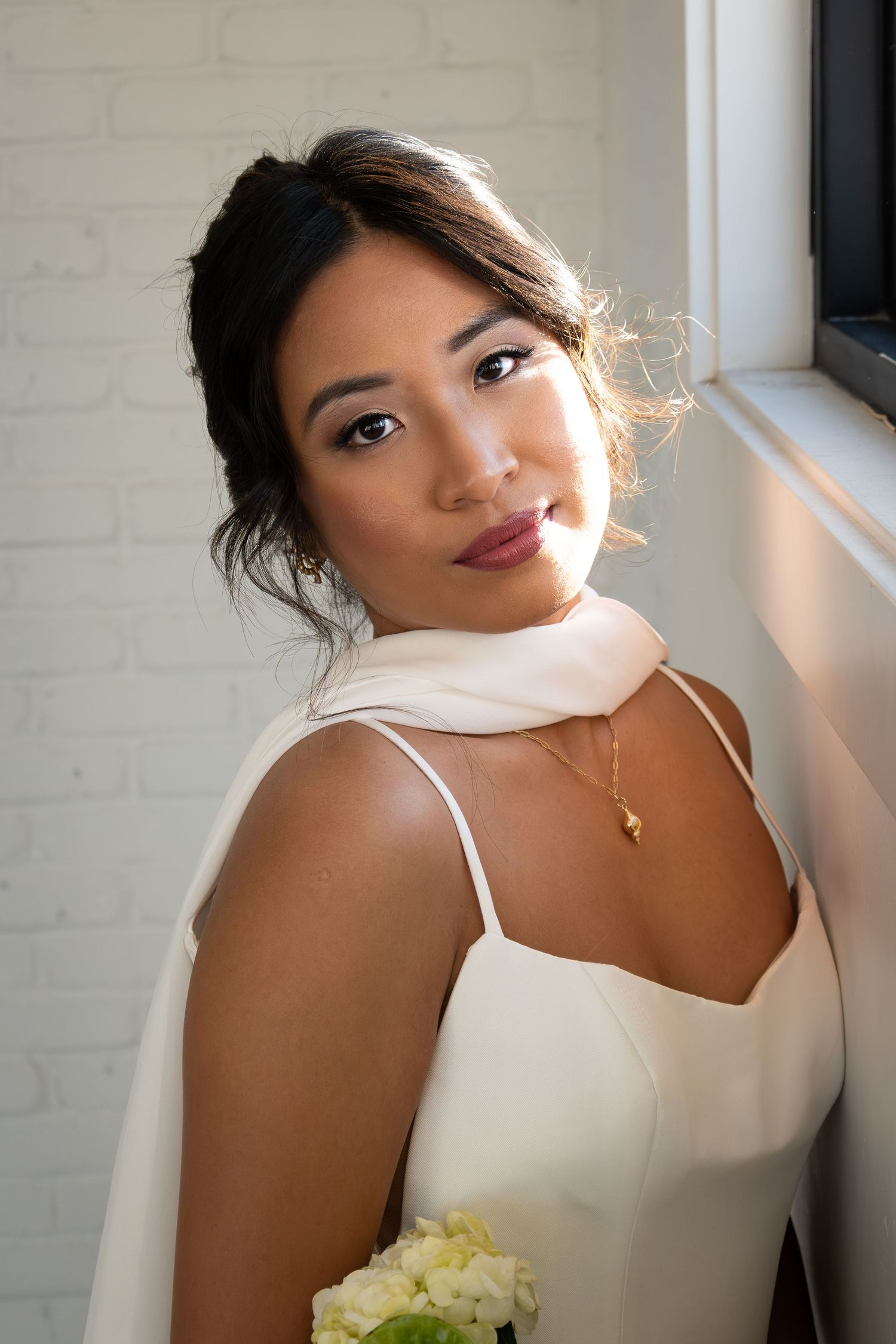 Elegant bride in white slip dress holding white flowers, posing by a window with soft natural light.
