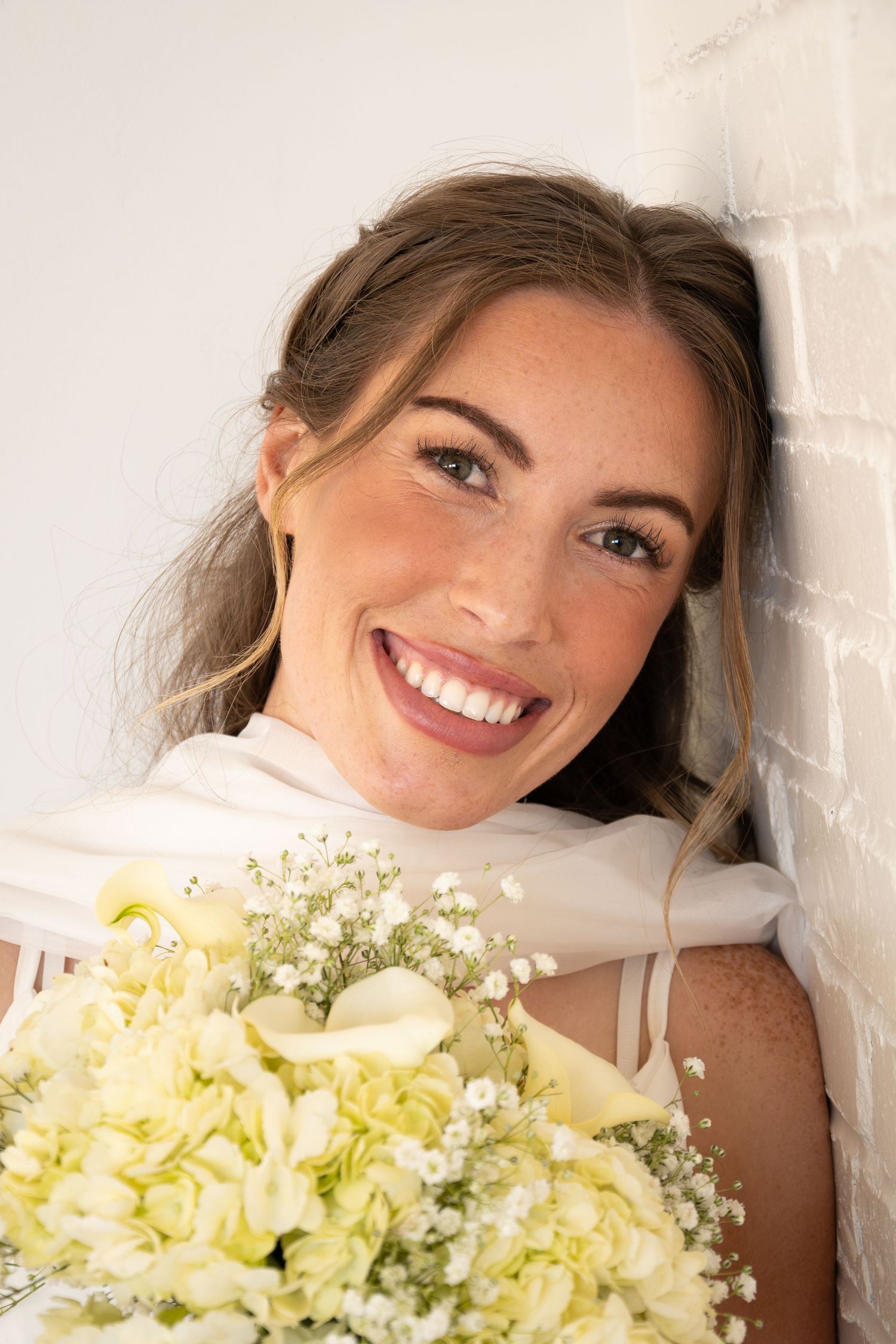 Happy bride with freckles leans against white brick wall holding a bouquet of white hydrangeas and calla lilies.