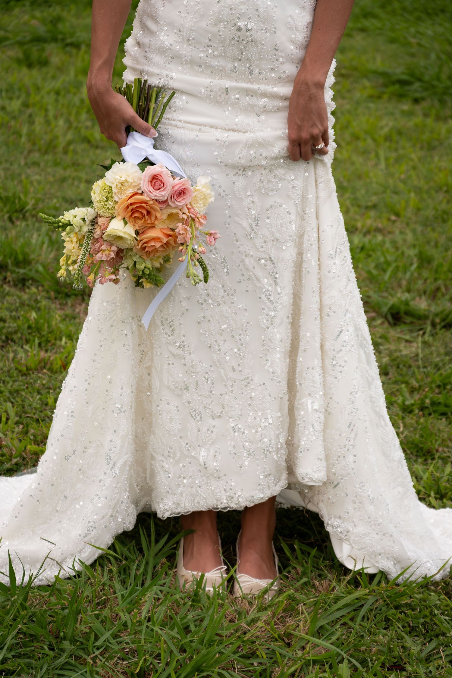 Bride in beaded white wedding gown holding peach and pink rose bouquet with white ribbon, standing on green grass.