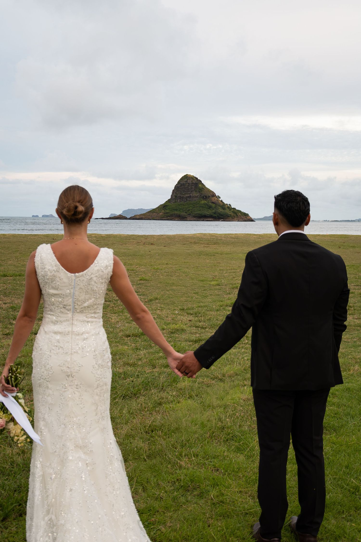 Bride and groom holding hands facing Chinaman's Hat island in Hawaii on their wedding day.