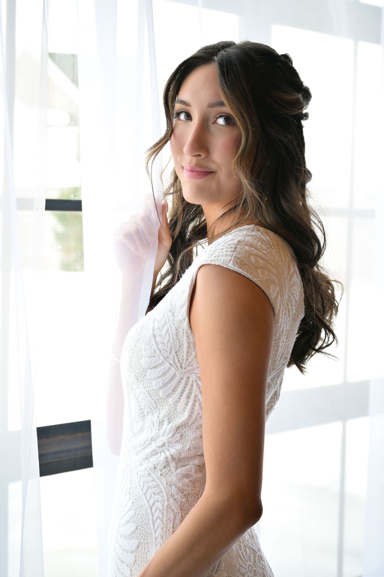 A bride in a white lace dress poses by a window with natural lighting.