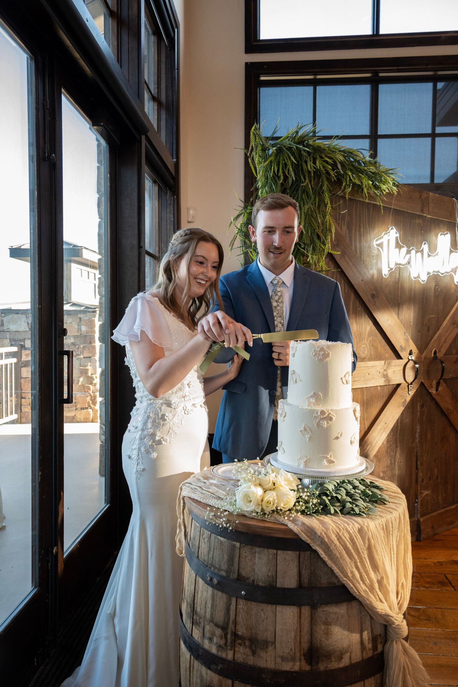 A wedding couple poses with an elegant white tiered cake displayed on a rustic bourbon barrel.