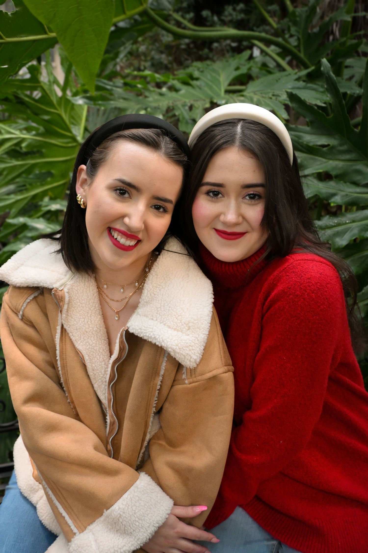 Two friends in winter coats smiling warmly while posing together against green foliage.