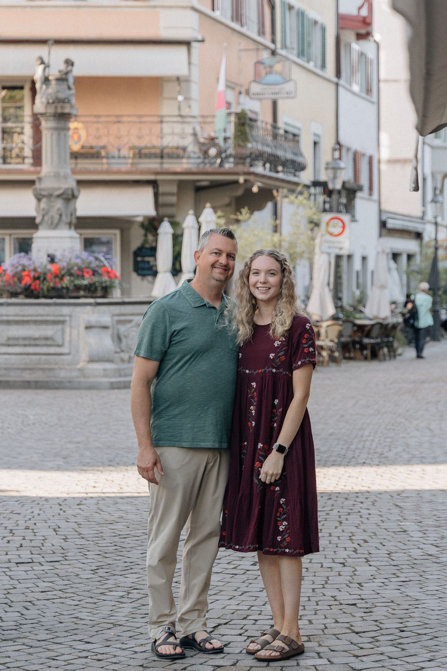 Two people standing together on cobblestone plaza with ornate stone column and buildings.