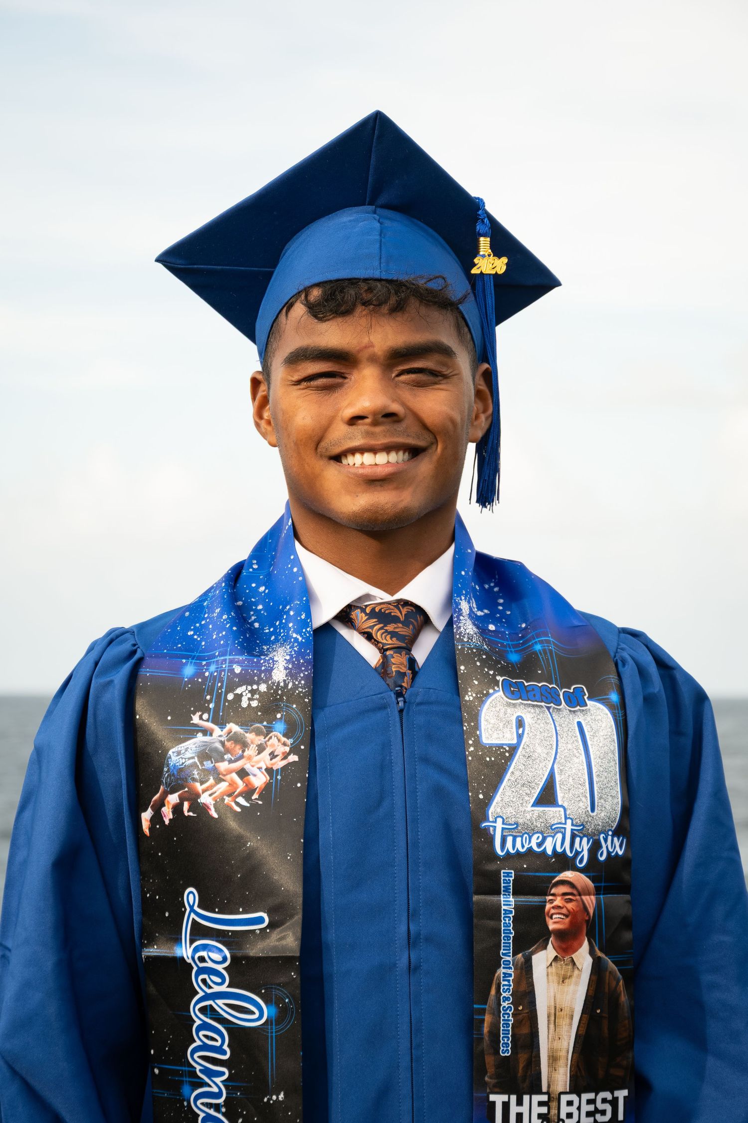 Smiling young man in blue graduation cap and gown with decorative stole outdoors on graduation day.