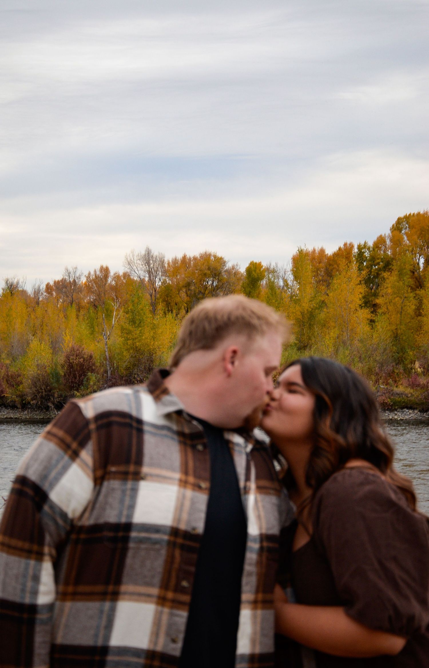 A couple shares a romantic kiss against an autumn backdrop of golden trees near a river.