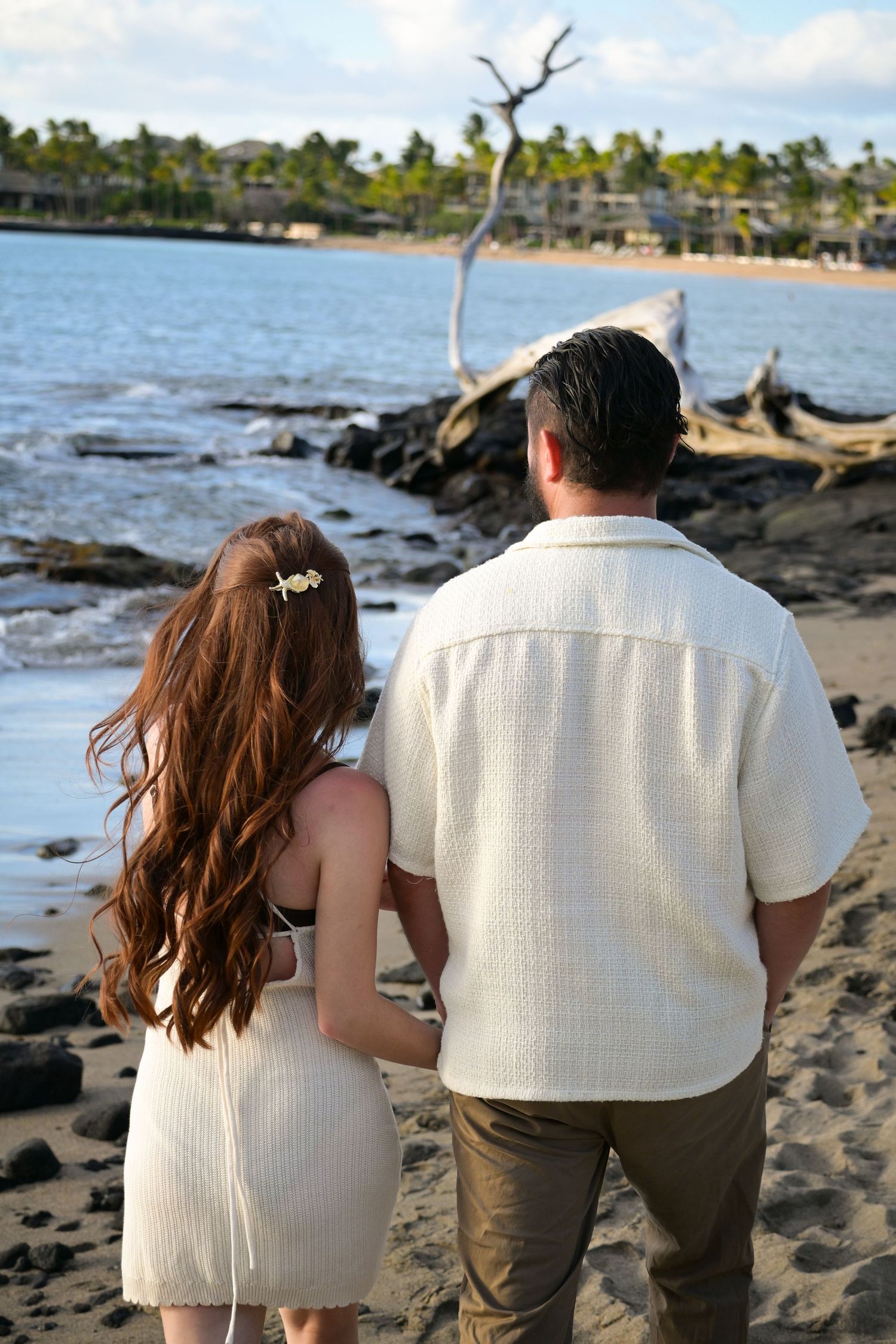 Couple stands together at edge of rocky Hawaiian beach with palm trees and ocean view in matching white outfits.