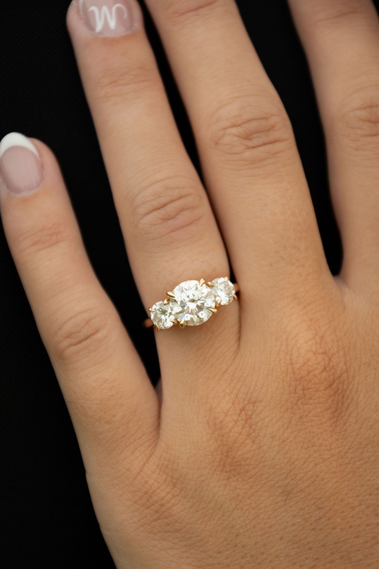 Three-stone diamond engagement ring in yellow gold setting worn on a woman's finger with French manicure.