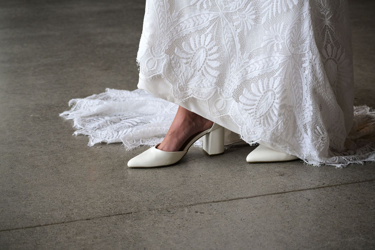 White lace wedding dress hem and bridal shoes on concrete floor.