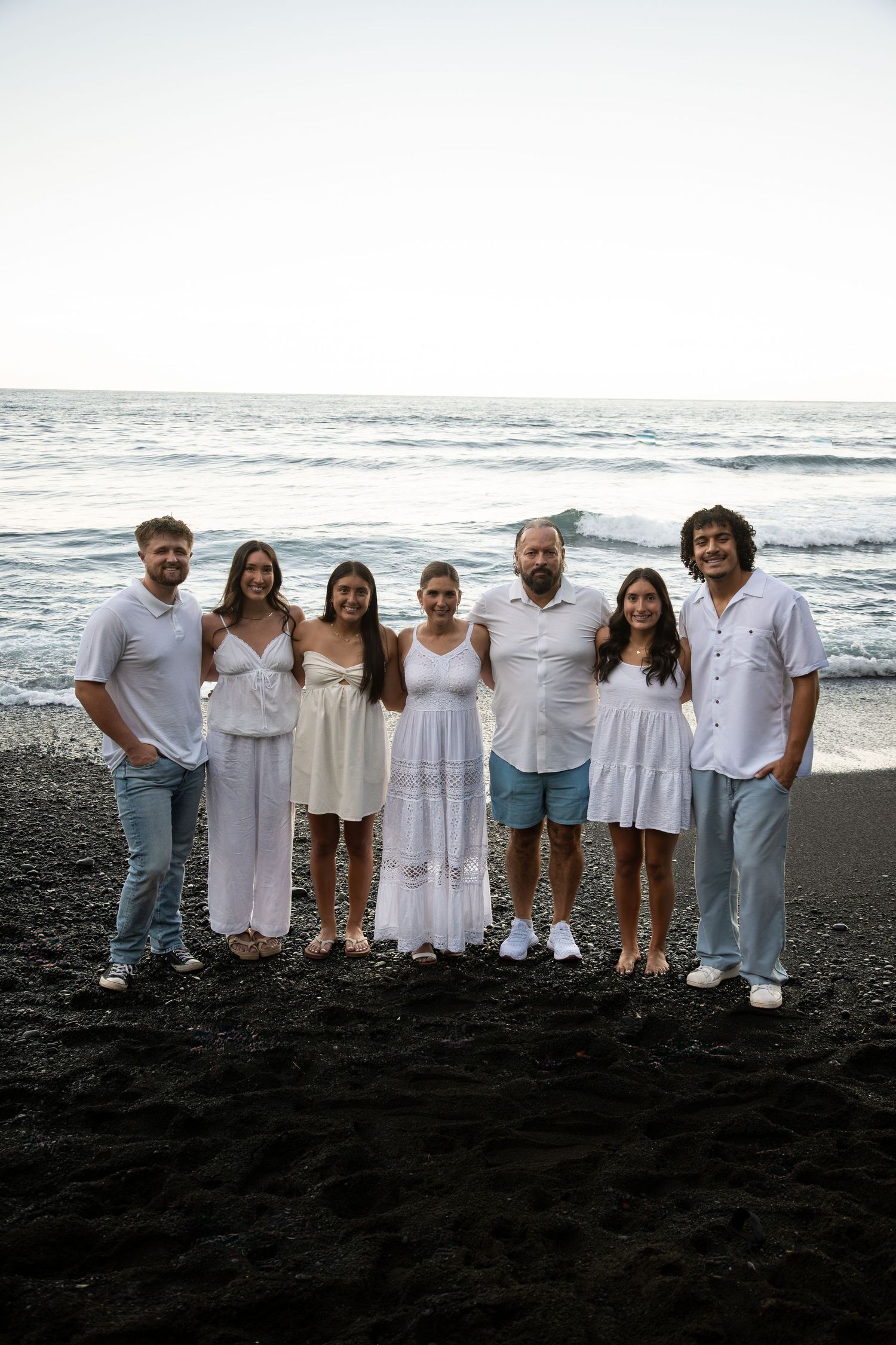 Family group wearing white clothing poses together on black sand beach with ocean waves in background.
