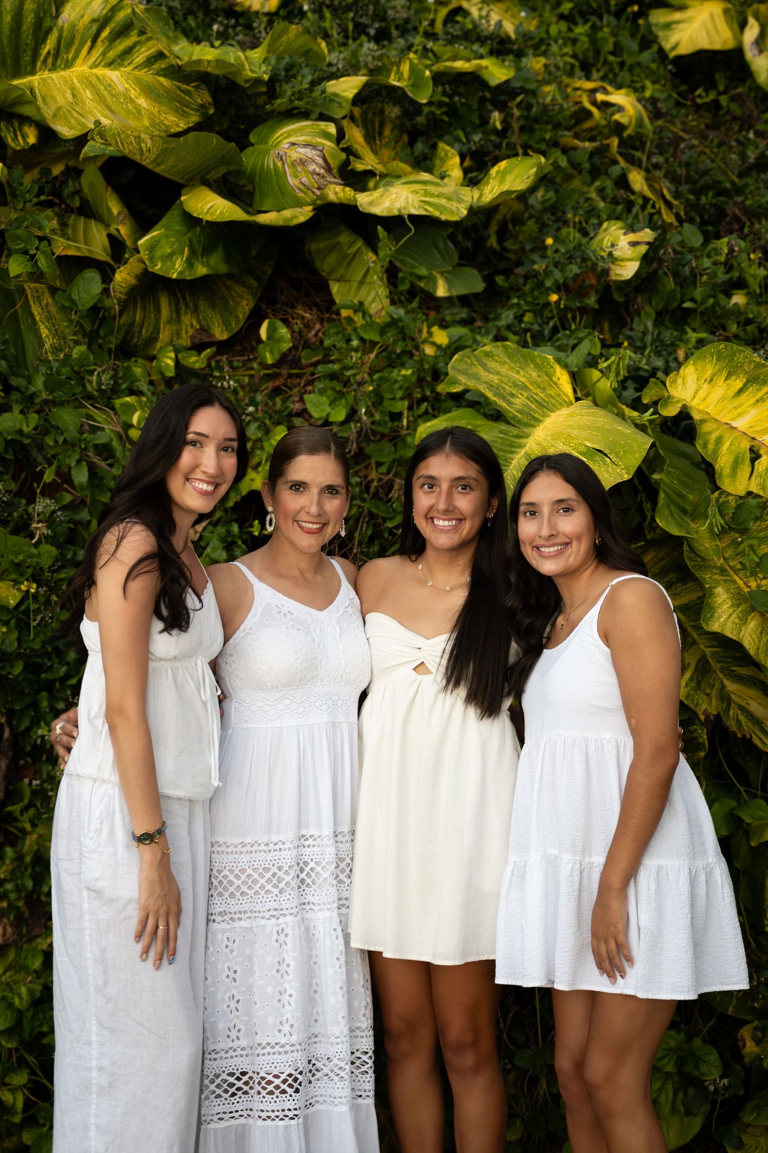 A group in white dresses poses together against a backdrop of lush tropical foliage with large yellow-green leaves.