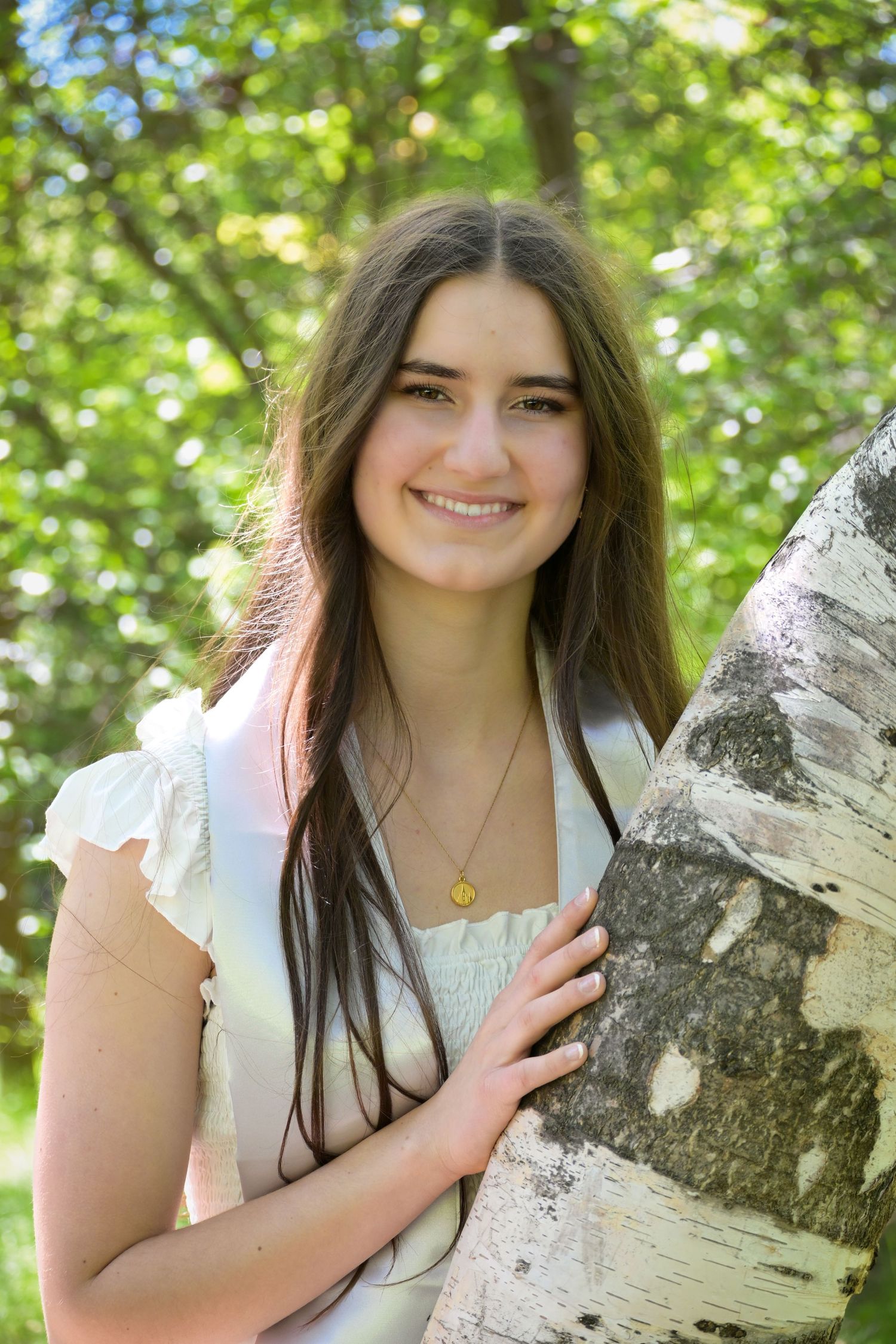 A person in a white top smiles while leaning against a birch tree trunk in a sunny outdoor setting.