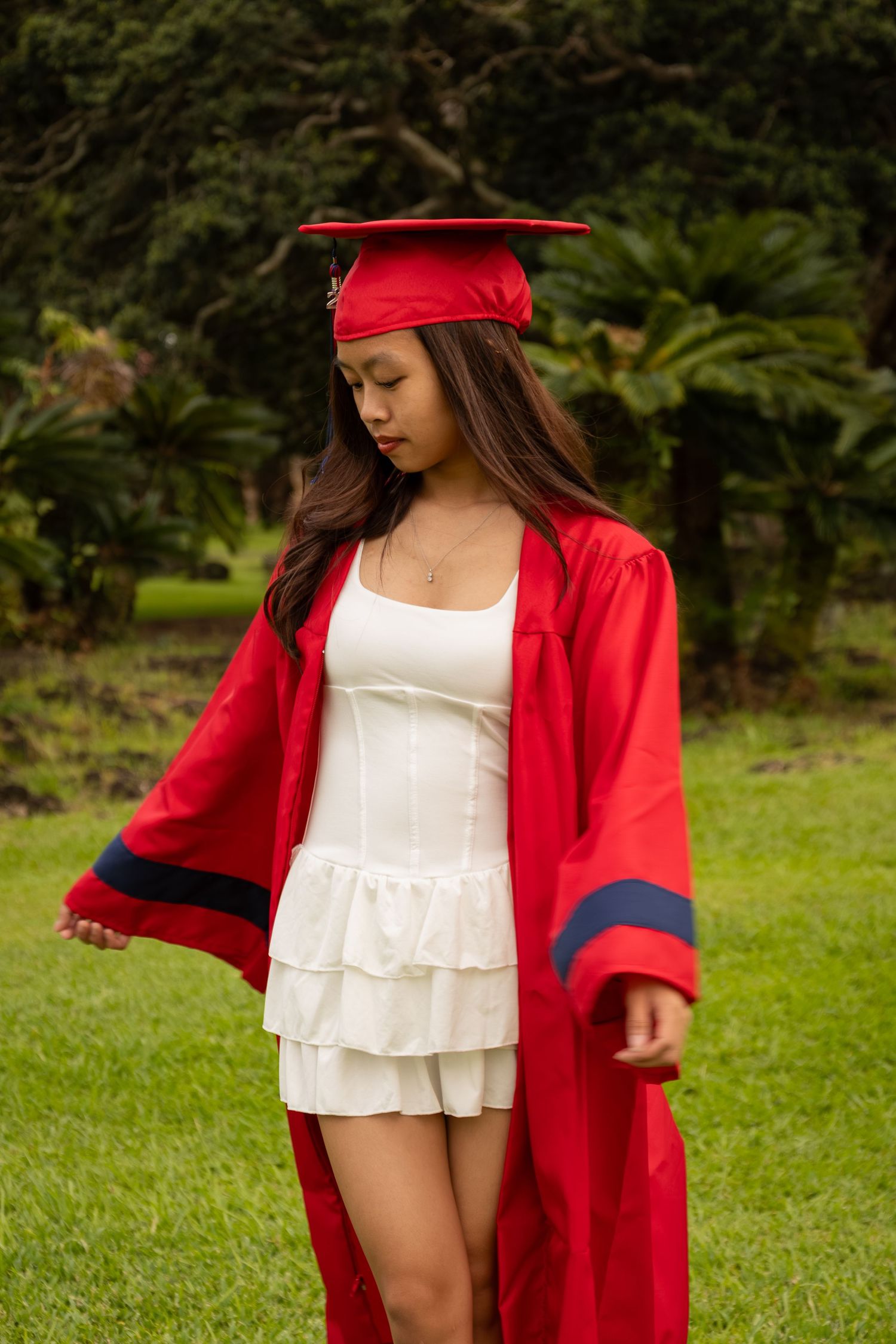 Young woman in red graduation cap and gown over white dress, posing outdoors on green grass with trees behind her.